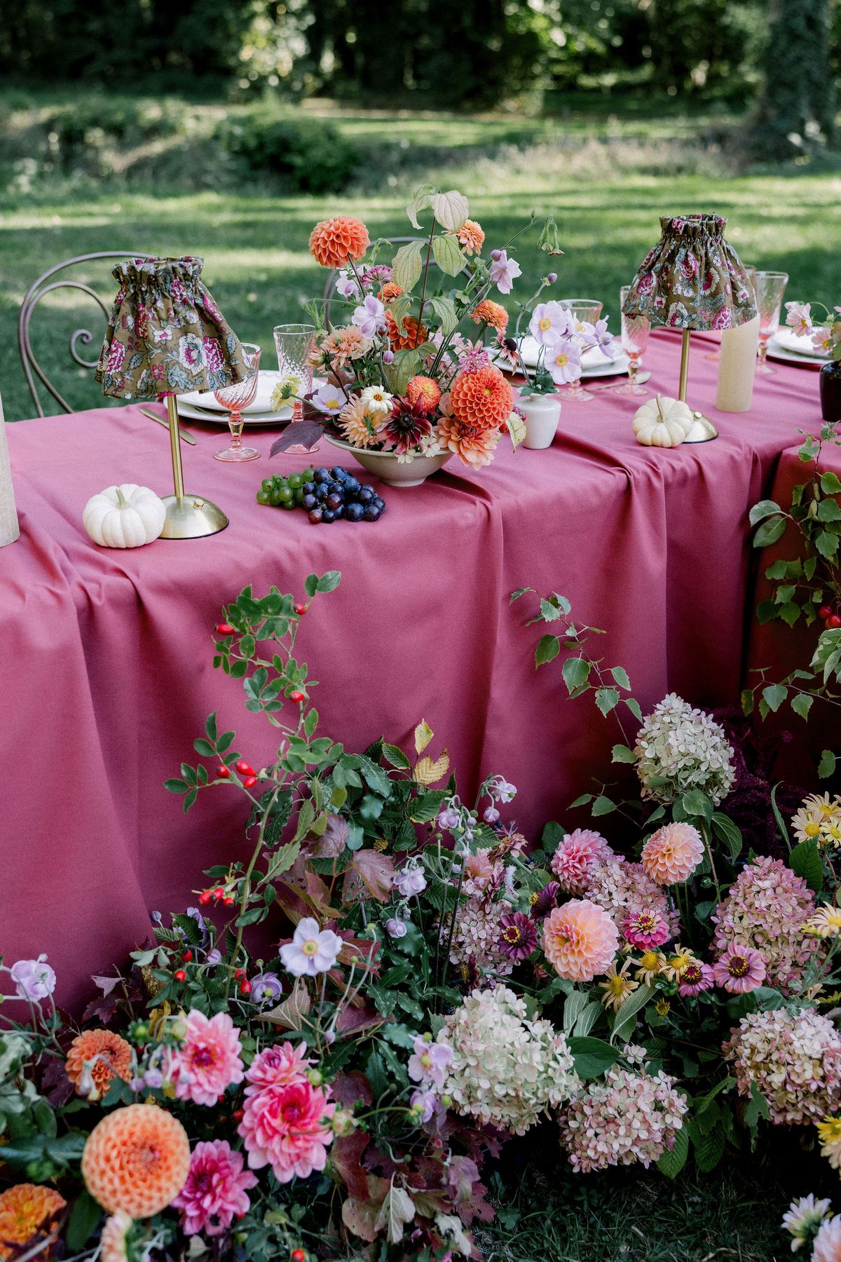 A table with a raspberry cloth holds an orange floral centrepiece, small lamps and grapes, with flowers massed on the grass below.