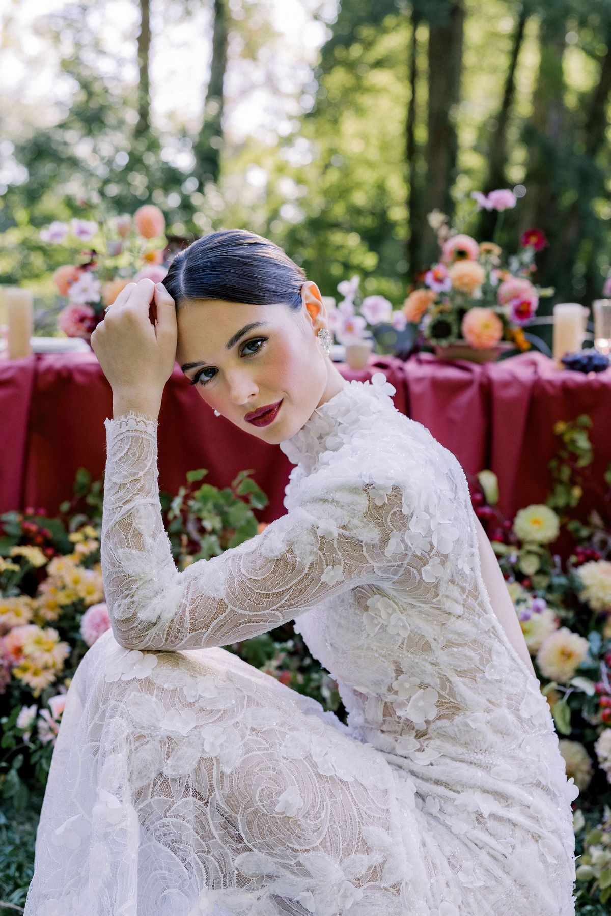 A bride in a lace gown with red lipstick rests her head on her hand, with a flower-laden table behind her.