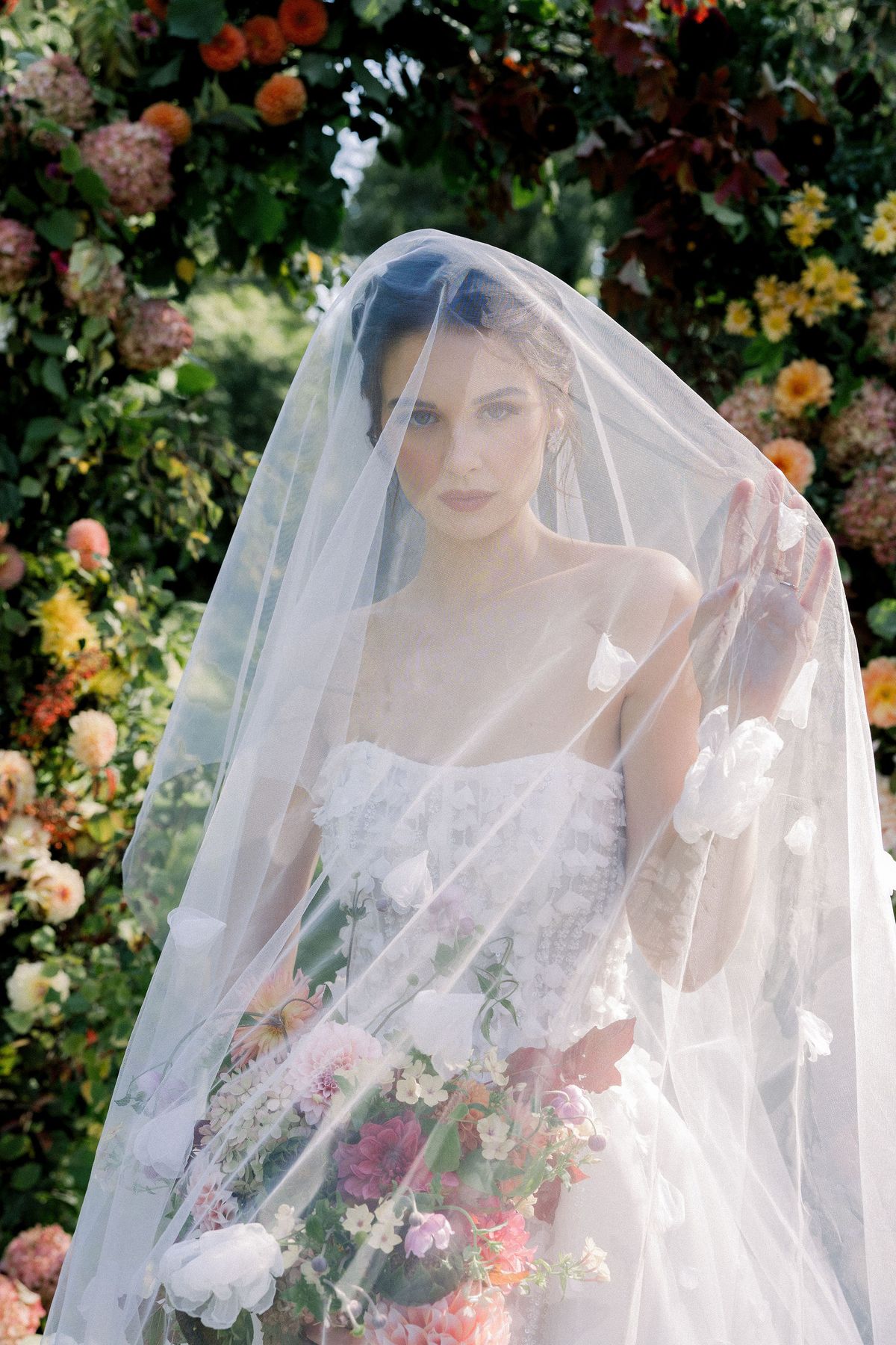 A bride stands with a sheer veil draped over her head and shoulders, holding flowers, in front of an arch of blooms.