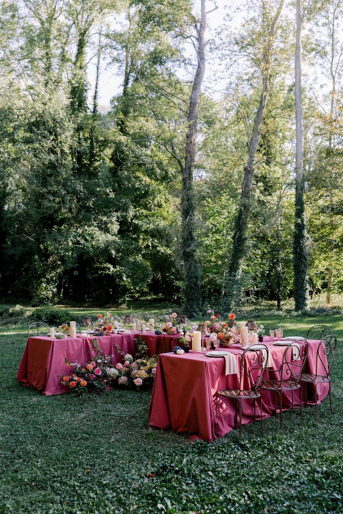 Burgundy-covered tables with flowers and chairs sit on a lawn in a clearing surrounded by slender trees.
