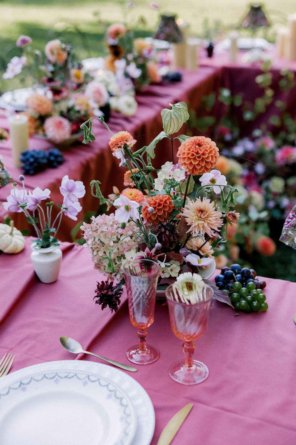 A long table with a burgundy cloth is set with white plates, pink glassware, grapes and a centrepiece of orange and pink flowers.