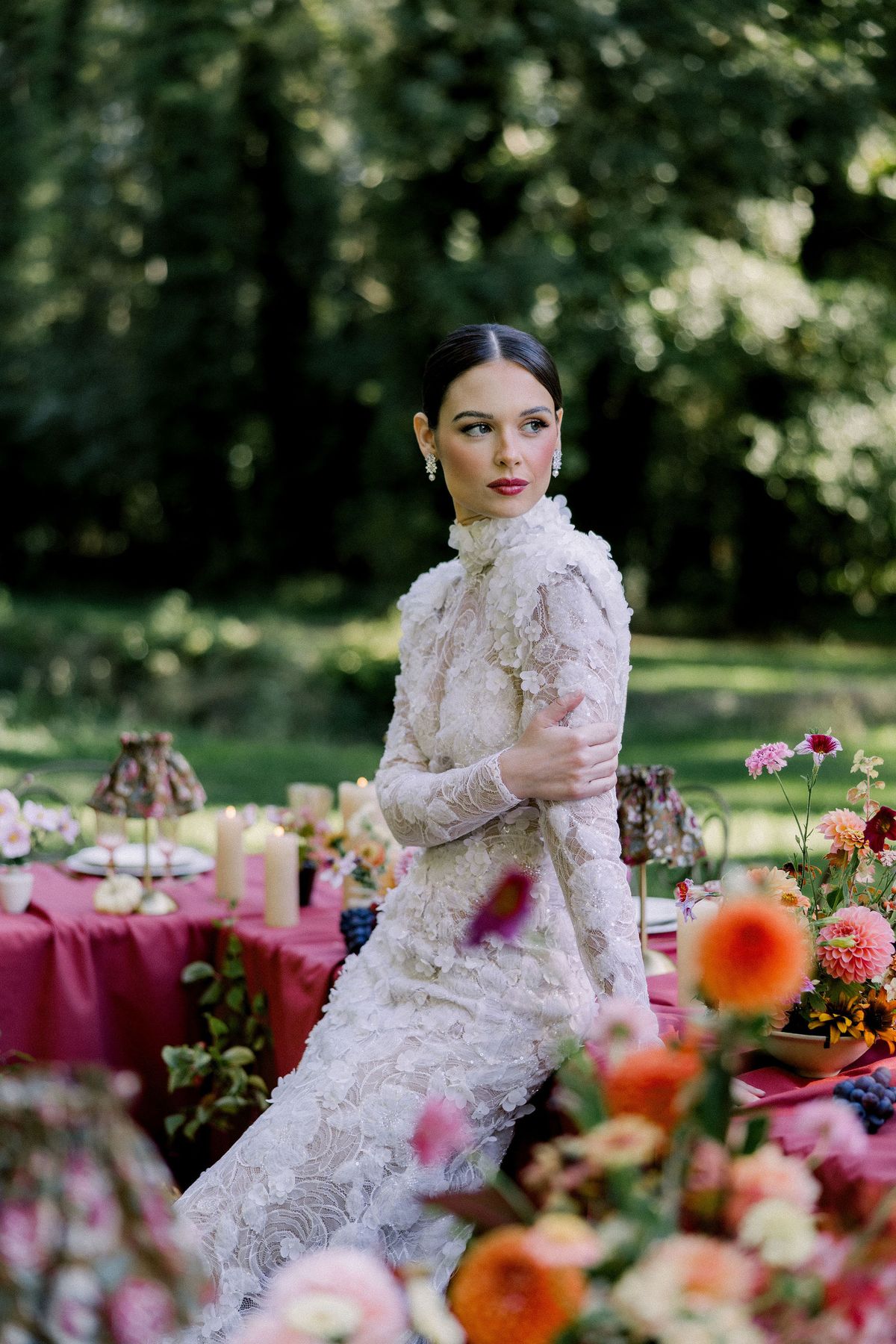 A bride in a lace gown with a high neckline sits near a table and bright orange flowers, glancing to the side.