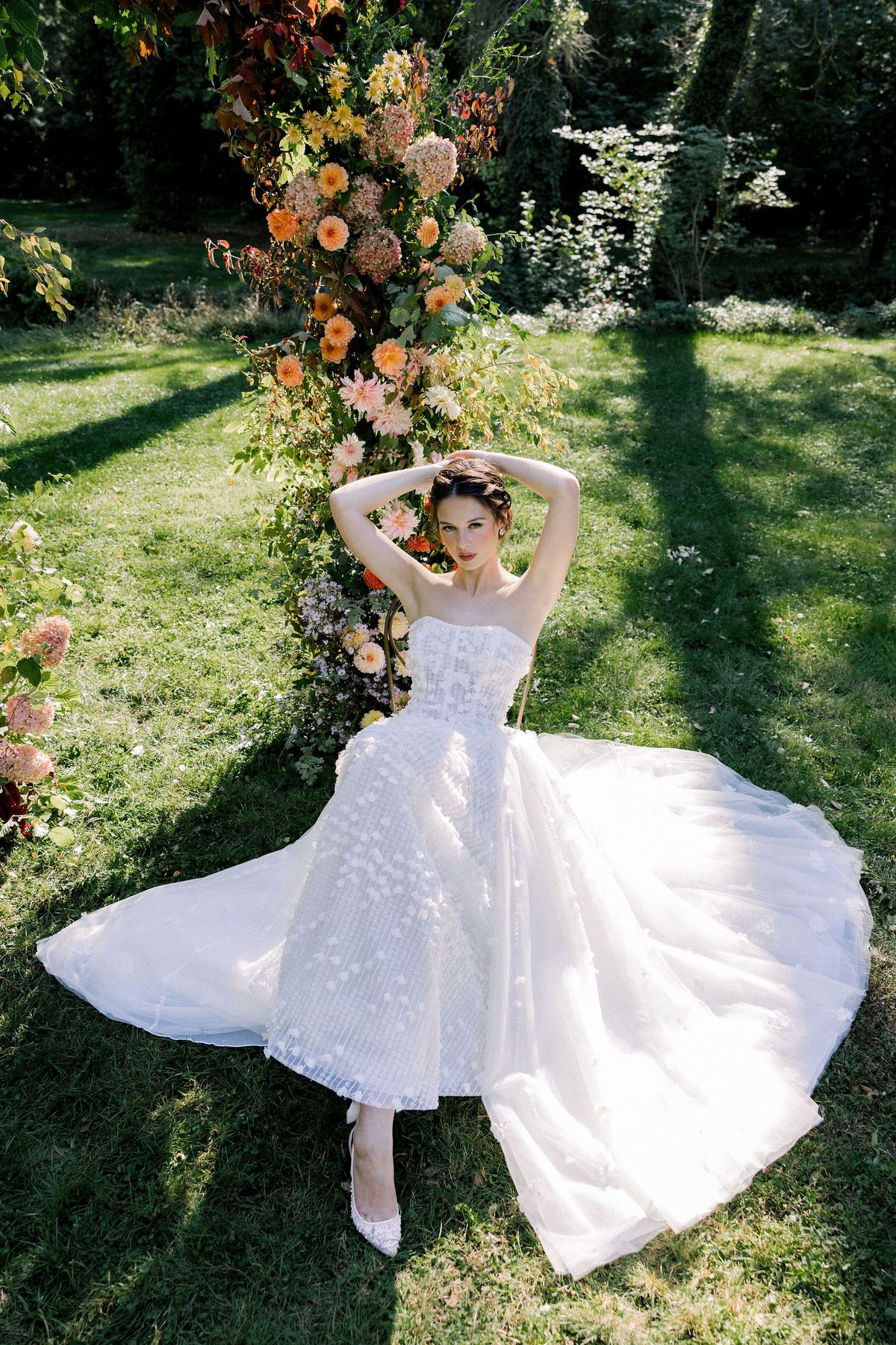 A bride in a strapless gown sits on a sunlit lawn with her arms raised, her full skirt spread out before a floral arch.