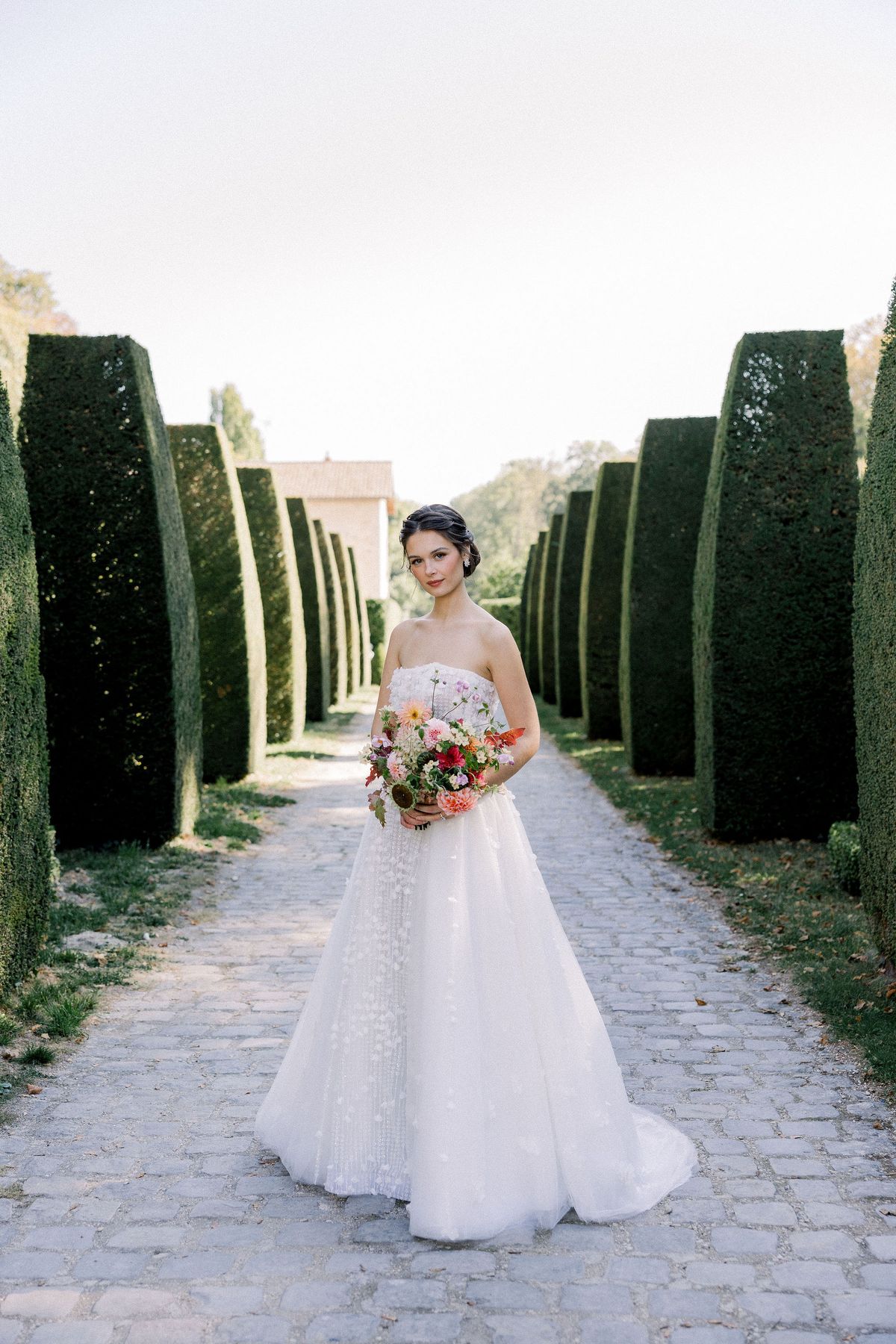 The bride in a strapless gown holds a bouquet on a cobbled path lined with tall clipped hedges.