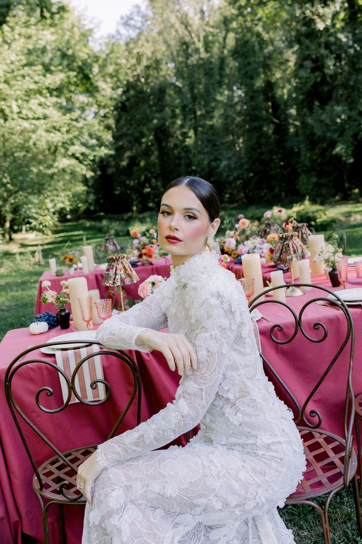A bride in a long-sleeved lace gown sits on an ornate metal chair beside a table with a burgundy cloth and candles.