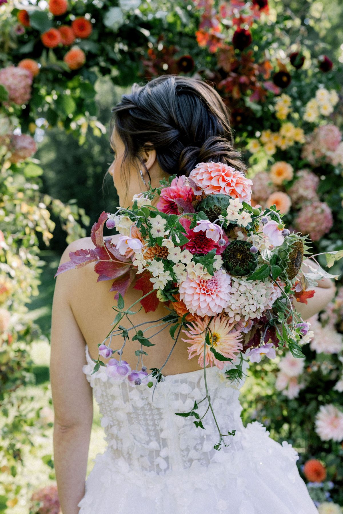 A bride with a dark updo faces away, holding a bouquet of pink, red and orange flowers in front of a floral arch.
