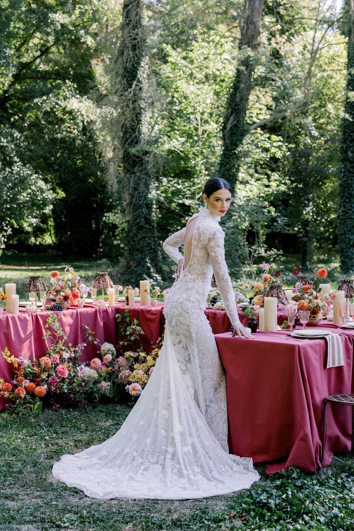 A bride in a long lace gown stands with her back partly to the camera beside a table dressed in raspberry and flowers.