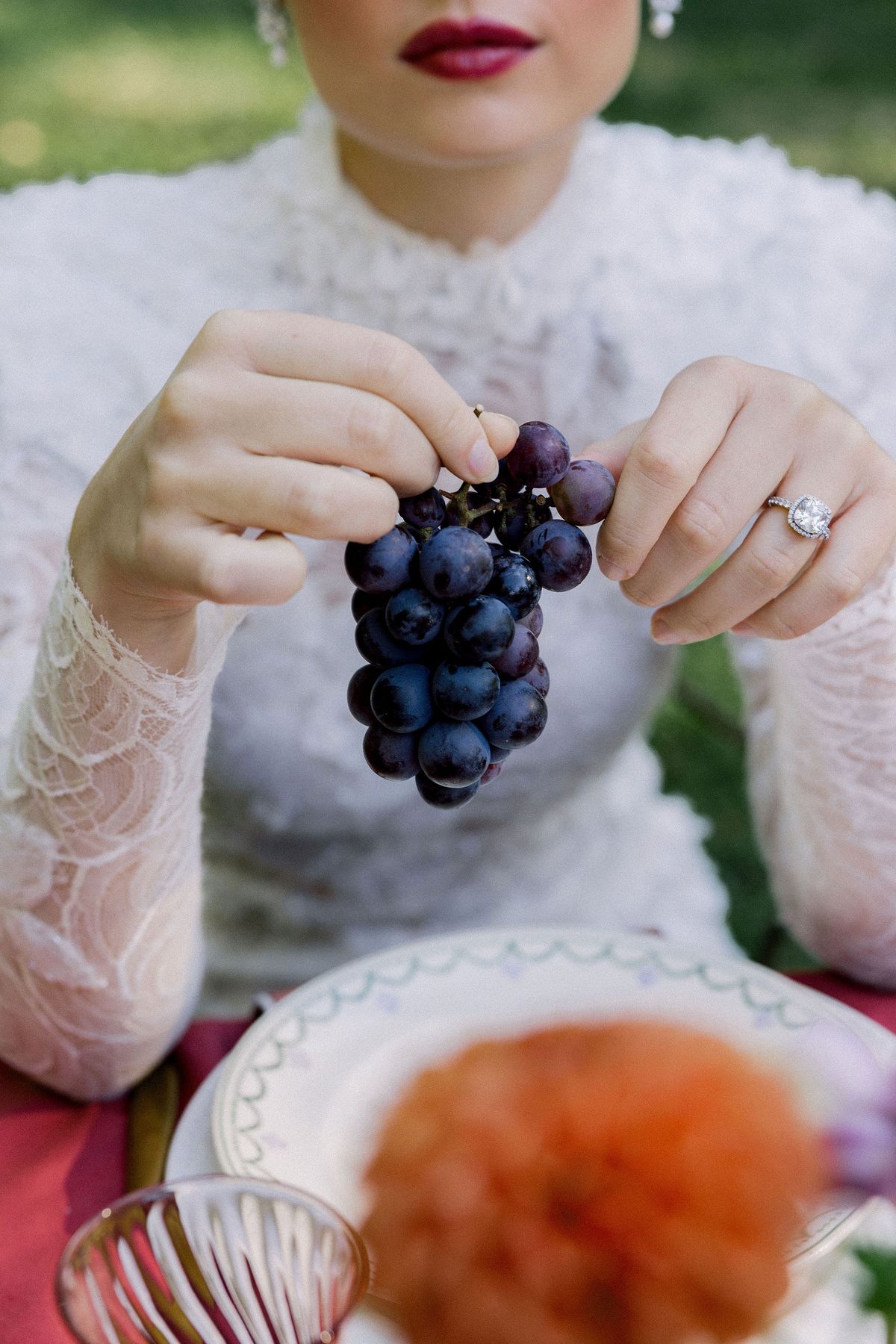 A bride in a lace gown holds a cluster of dark grapes above a decorated plate, wearing a ring.