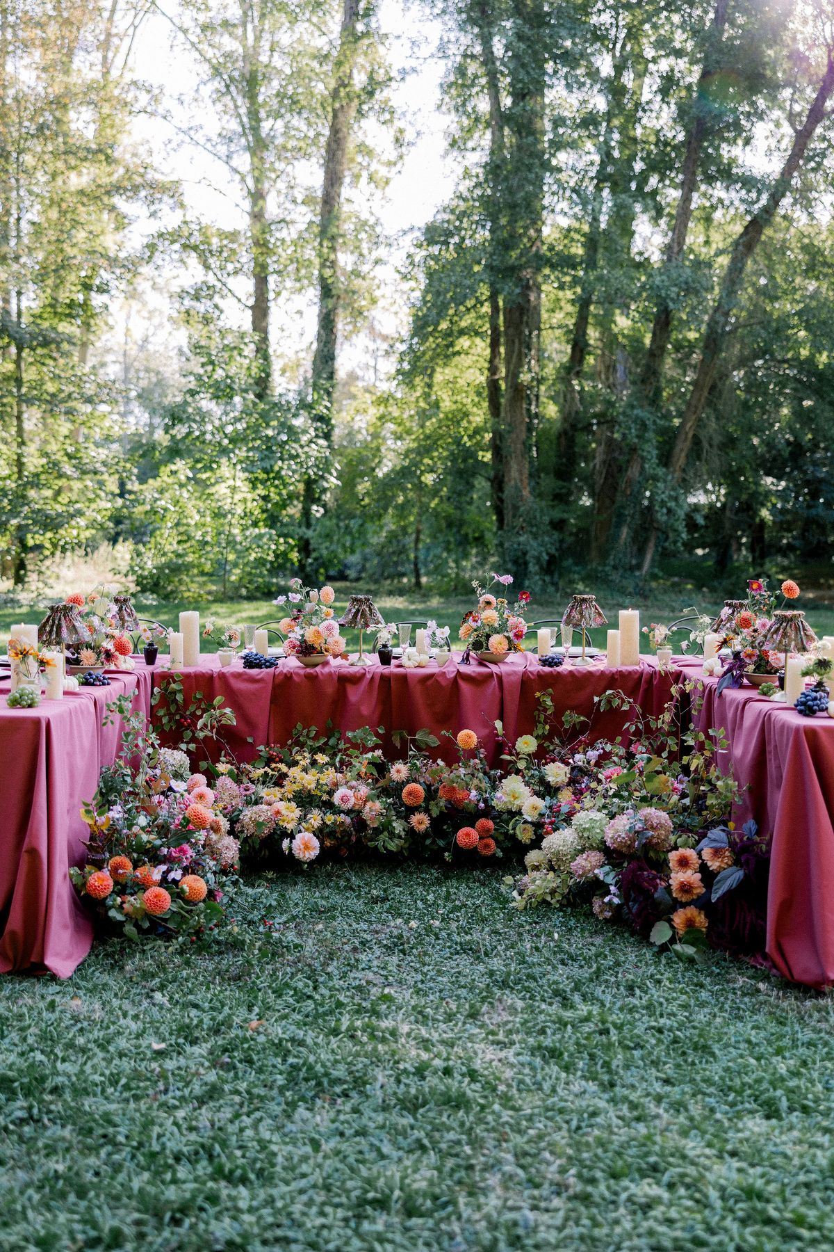 A burgundy-clothed table in a U shape edged with colourful flowers stands on grass below tall trees.