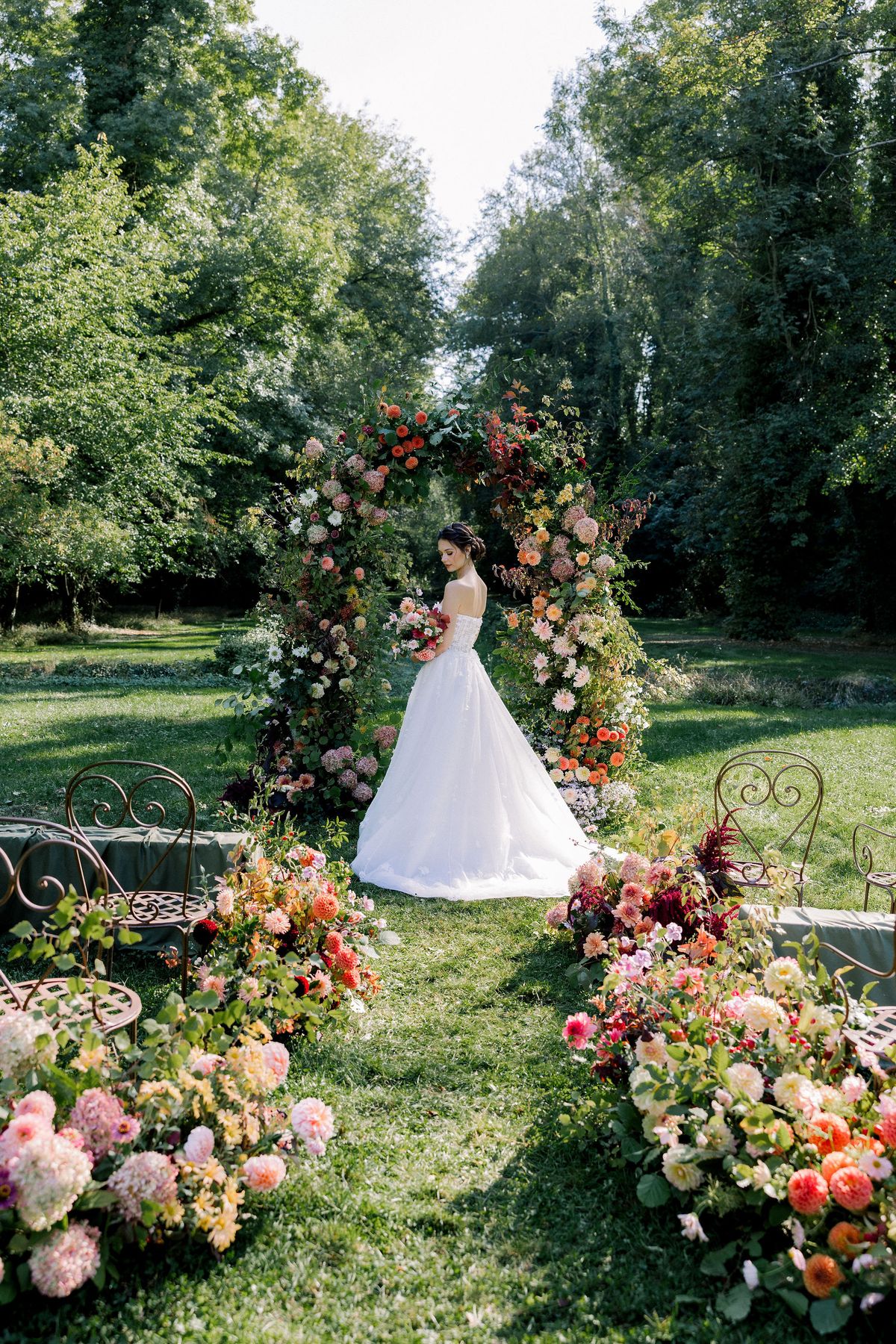 A bride in a white gown stands on the lawn in front of a tall floral arch, with flower borders and trees around.