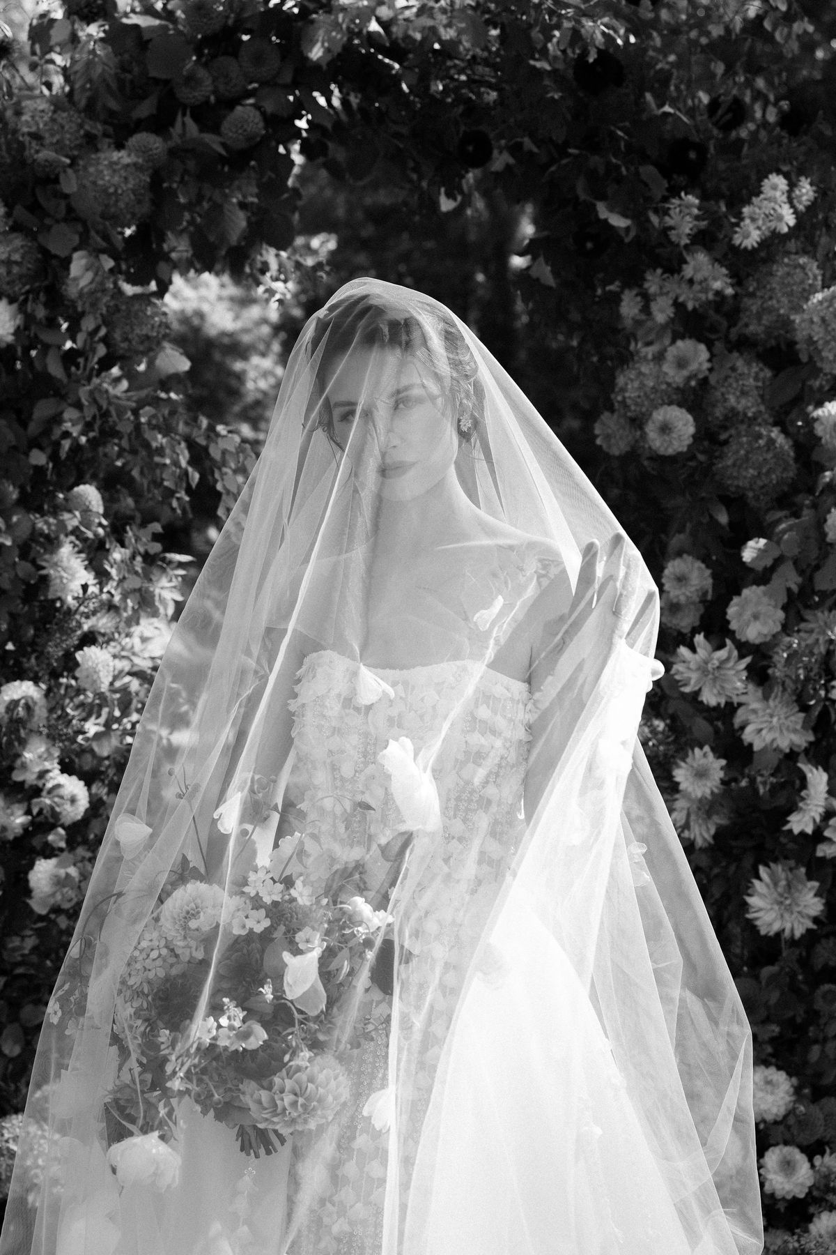 A black and white image of a bride with a sheer veil covering her head, framed by dark flowers behind.