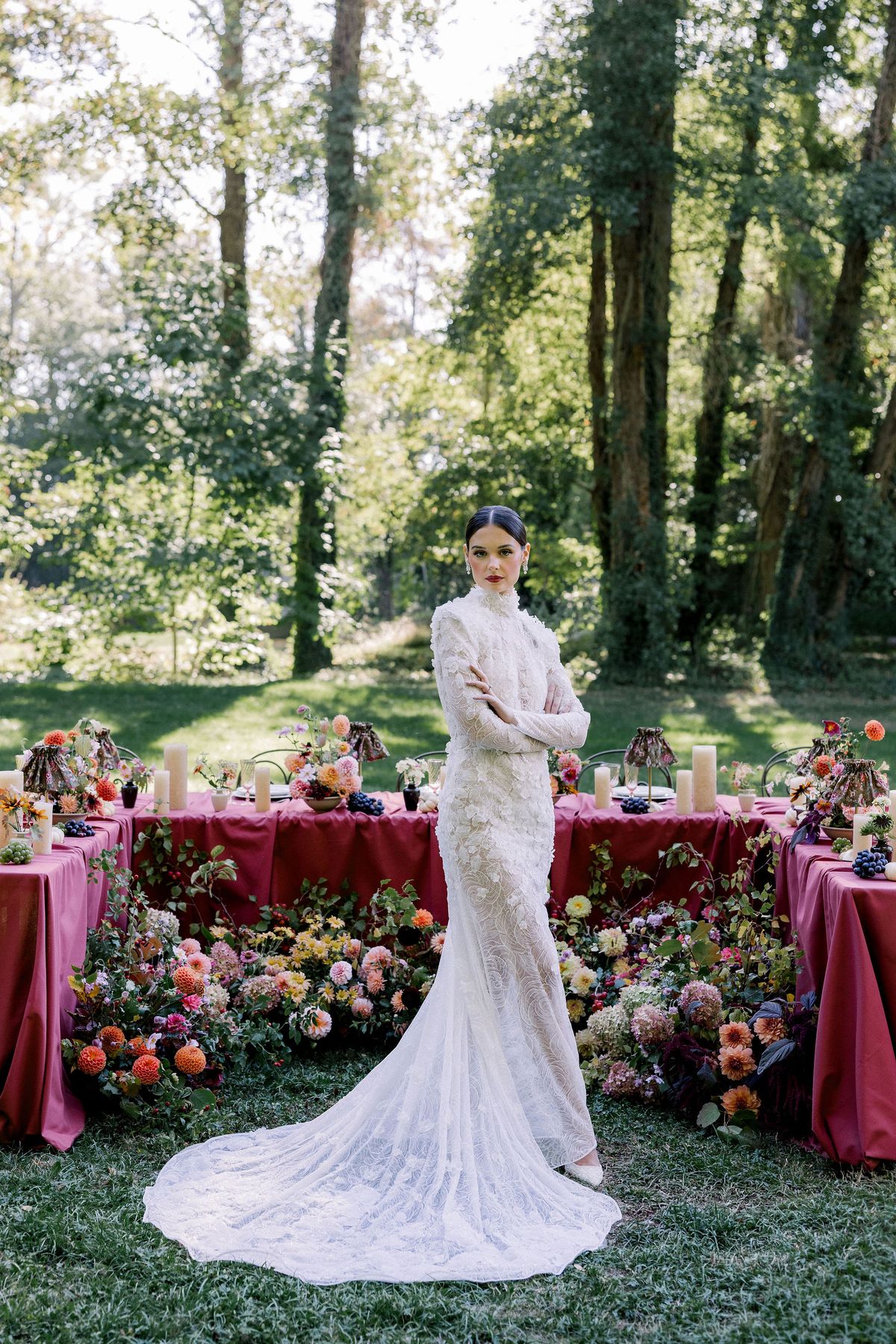The bride in a fitted lace gown with a long train stands with her arms folded in front of a burgundy table and flowers.