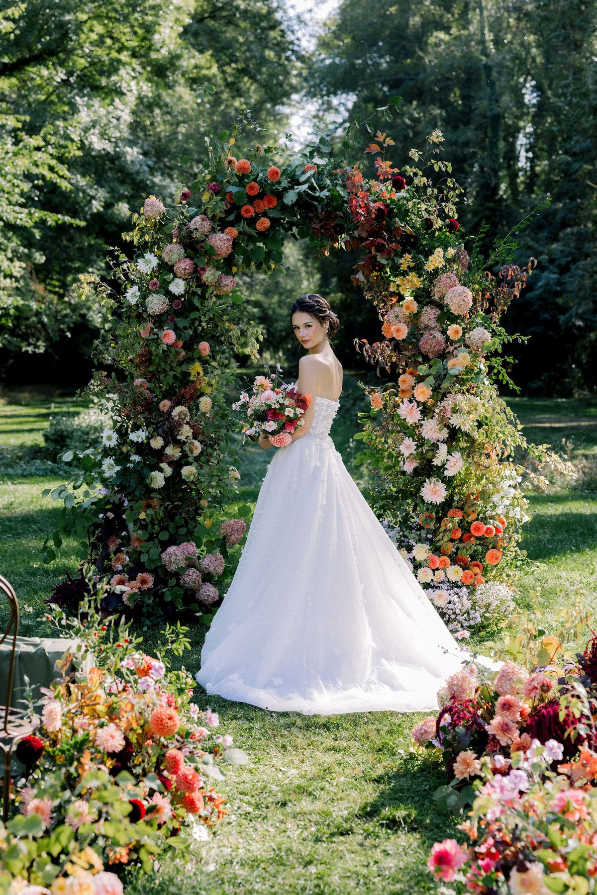 The bride in a full white skirt holds a bouquet and stands beneath a tall arch of orange, pink and red flowers.