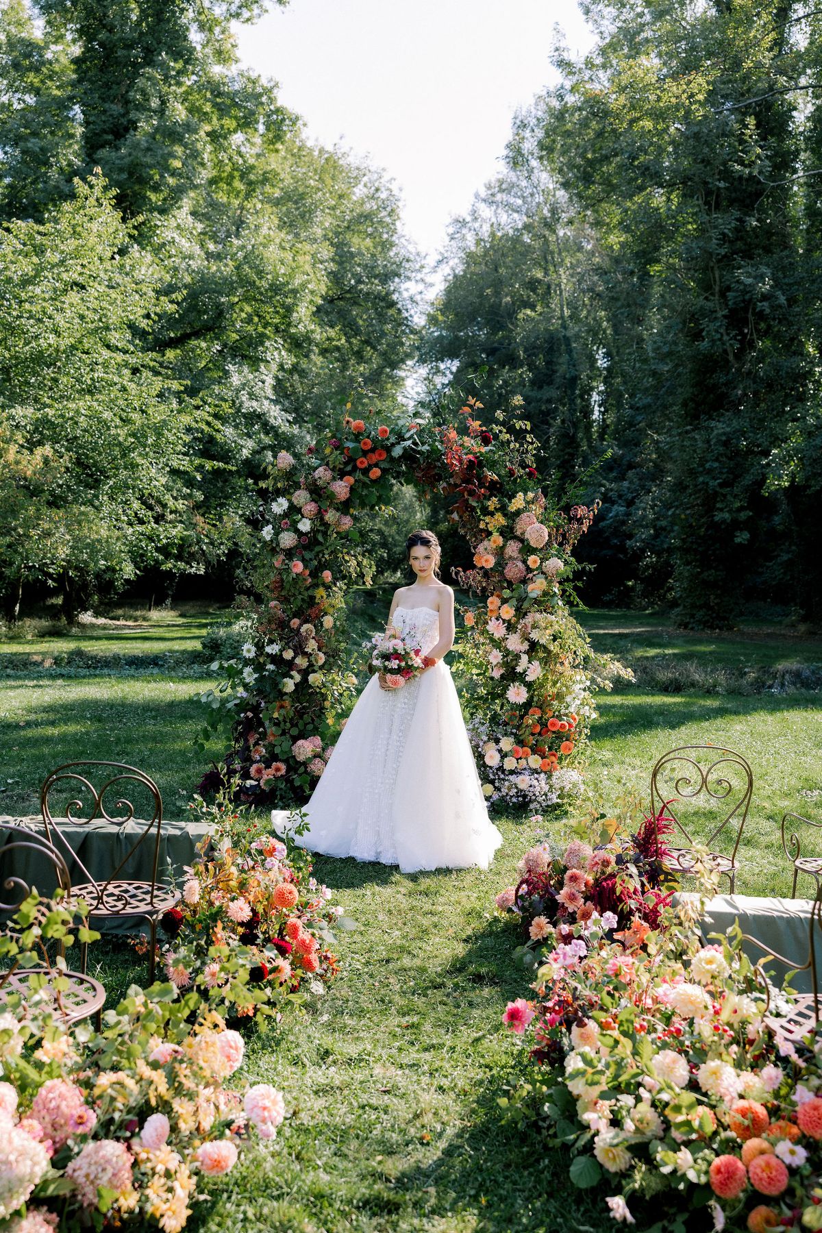 A bride in a full white skirt stands beneath a floral arch, with flower arrangements lining the grass and trees behind.