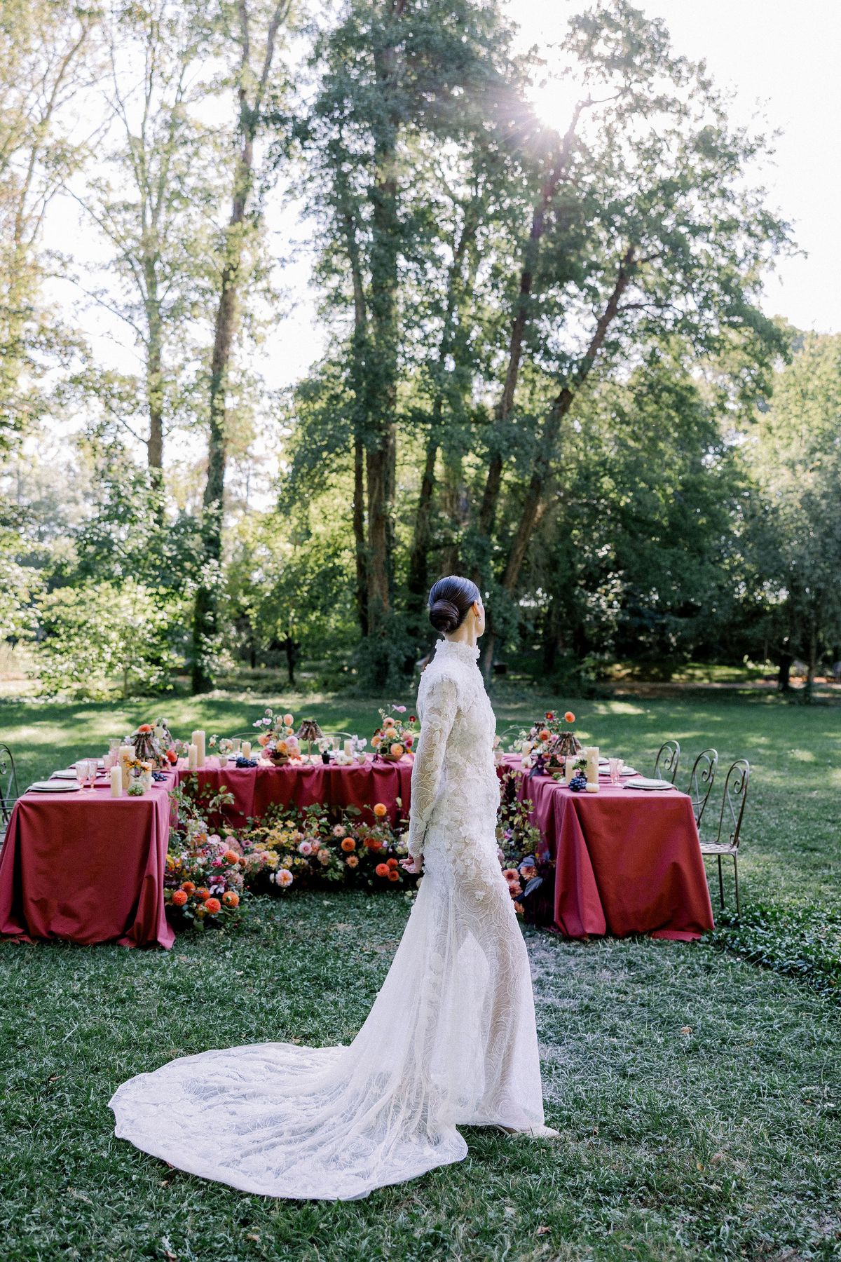 A bride in a long-sleeved lace gown with a train stands on grass, turned away towards a burgundy dining table among trees.