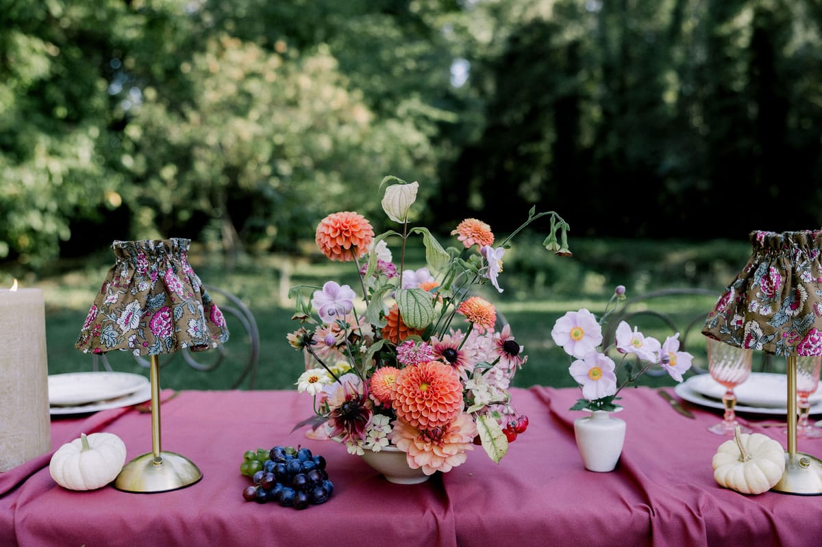 A burgundy-covered table holds a centrepiece of orange dahlias, small patterned lampshades, white pumpkins and dark grapes, with trees behind.
