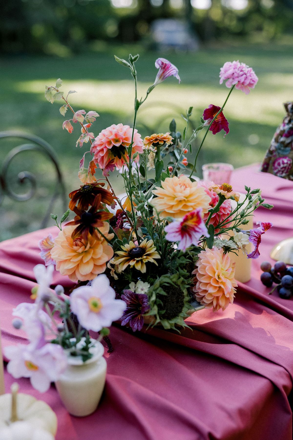 A loose arrangement of orange, pink and burgundy dahlias and small blooms sits on a burgundy tablecloth outdoors.