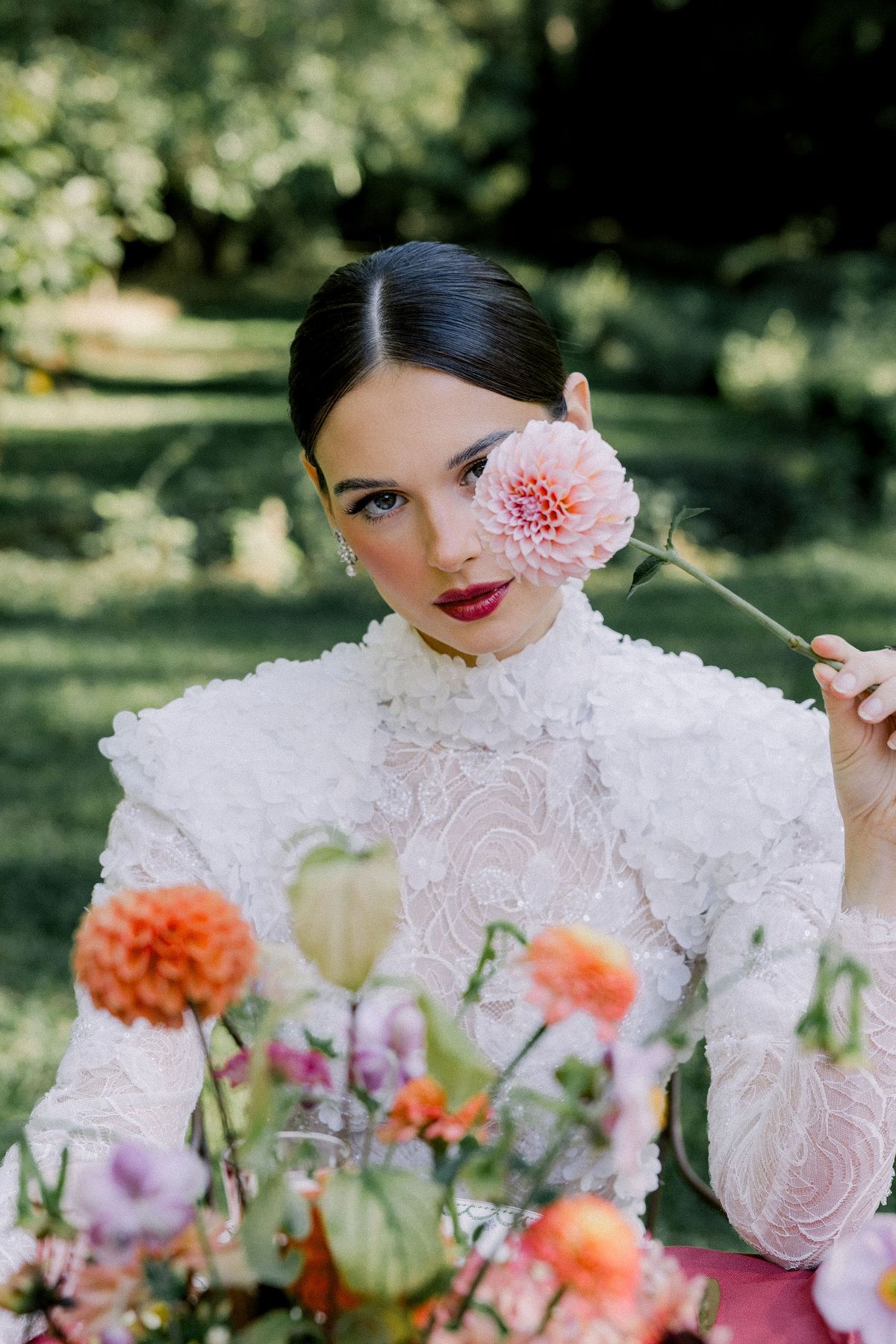 A bride in a high-necked white lace gown holds a coral dahlia in front of one eye, with colourful flowers in the foreground.