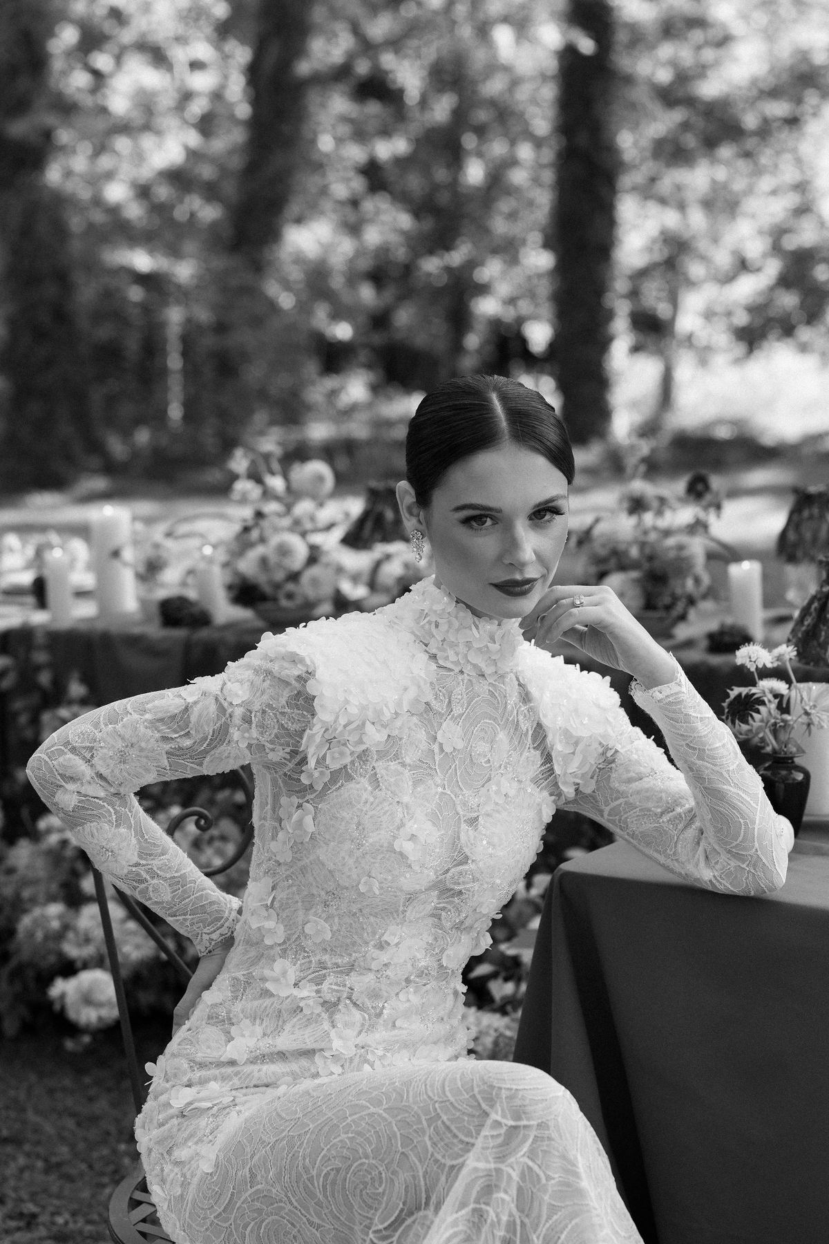 A black and white image of a bride in a high-necked lace gown sitting at a table, resting her chin on her hand.