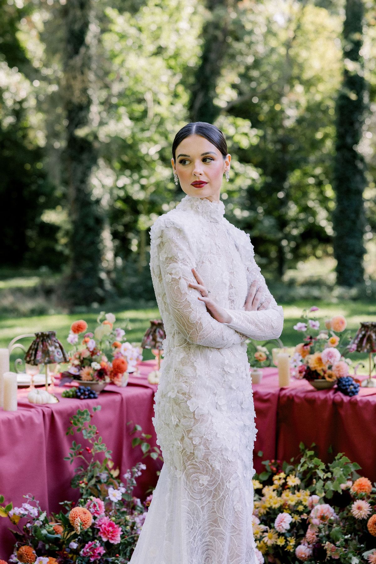 The bride with her hair swept back and red lips, wearing a high-necked lace gown, looks to the side with her arms crossed.
