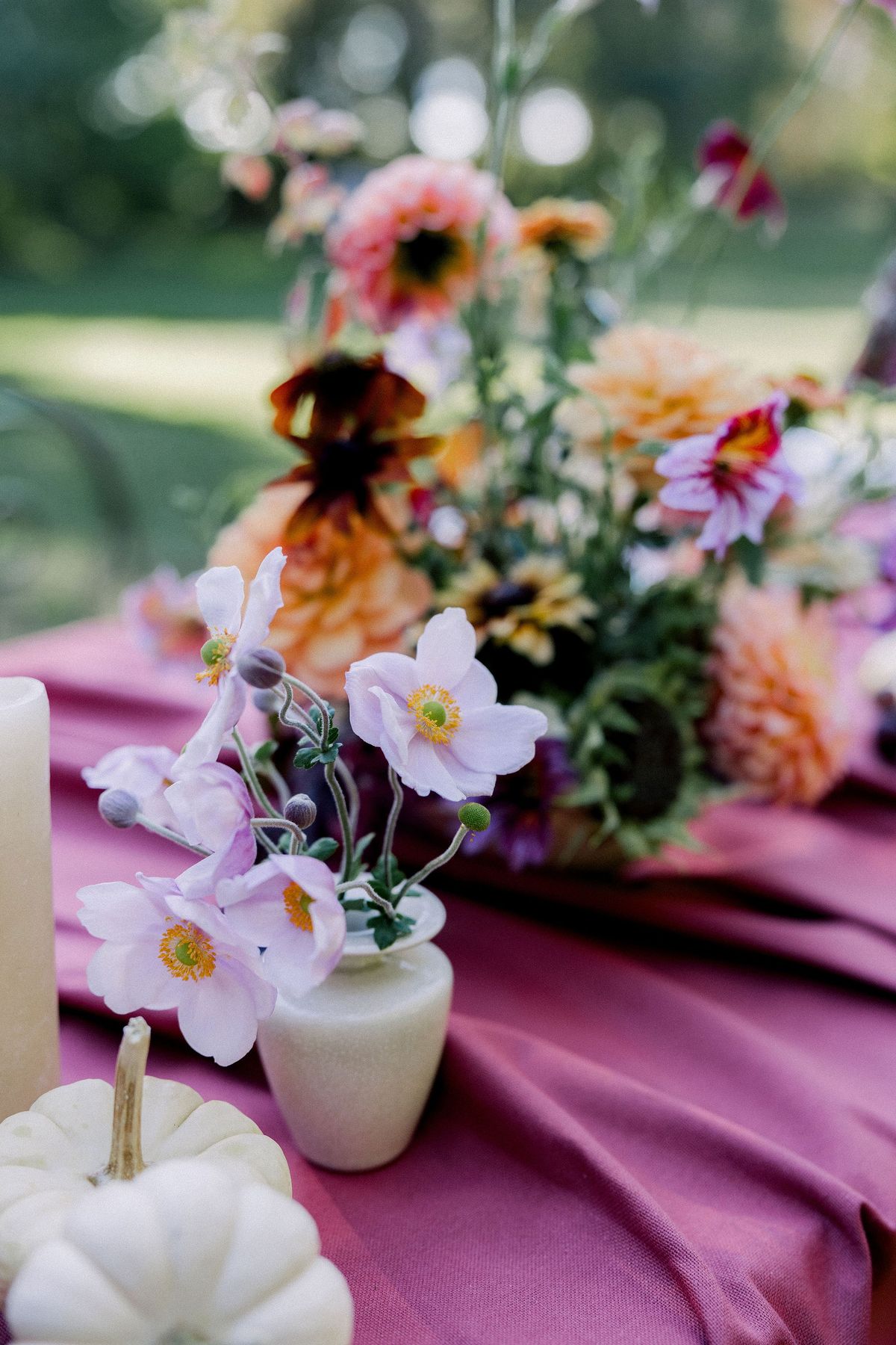 A vase of orange, pink and lilac flowers stands on a deep pink tablecloth beside a small white pumpkin.