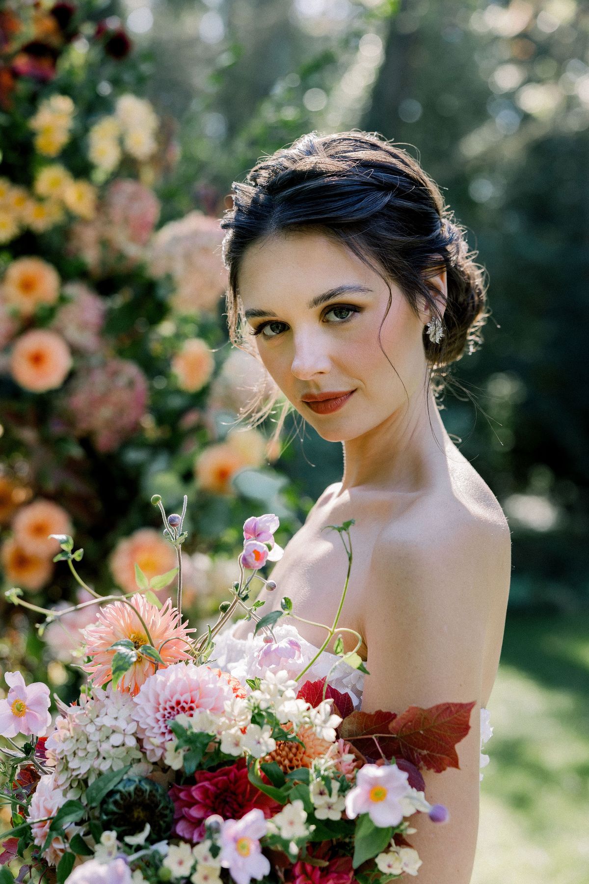 A bride with her hair pinned up looks towards the camera, holding a bouquet of pink, orange and red flowers in a sunlit garden.