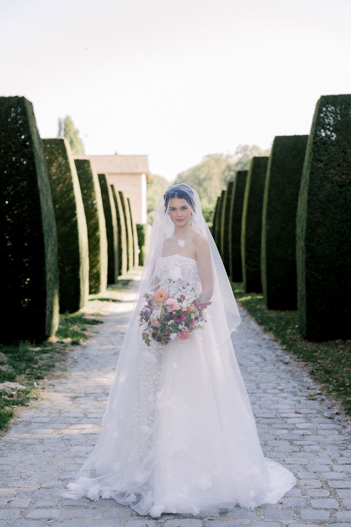 The bride in a veil holds a bouquet and stands on a cobbled path between tall clipped hedges leading to a building.