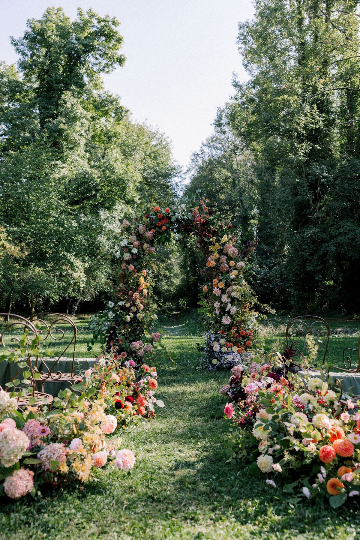 A tall floral arch stands on a lawn bordered by arrangements of pink and orange flowers, with tall trees on both sides.
