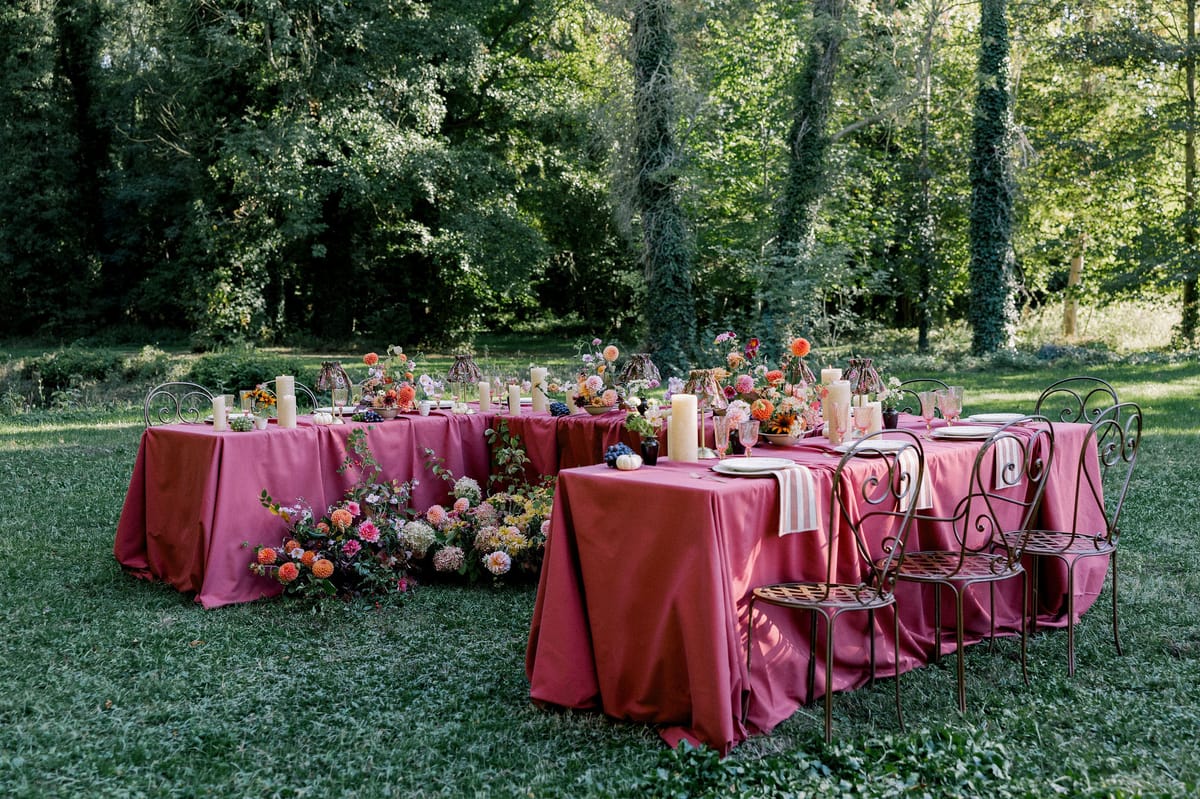 Burgundy-draped tables with flowers and candles stand on a lawn in front of tall trees, with wrought-iron chairs along one side.