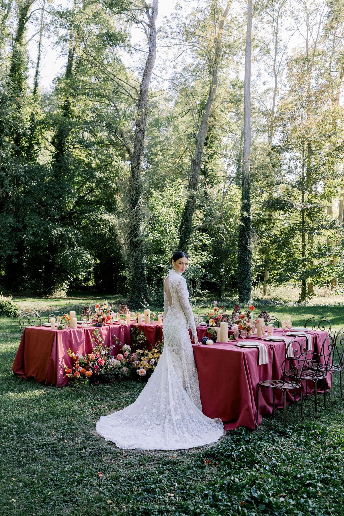 A bride in a lace gown with a long train stands beside a raspberry-covered table beneath tall trees.