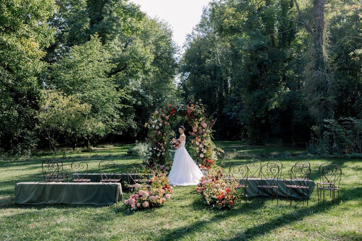 A wide view of a wooded lawn with a floral arch and chairs, with a small figure of the bride standing beneath it.