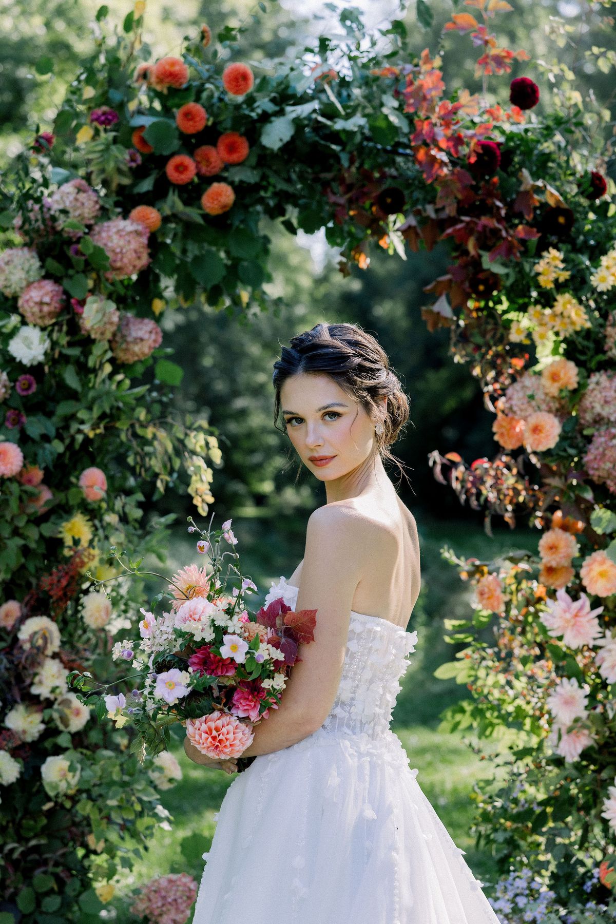 A bride in a strapless gown holds a bouquet and glances over her shoulder beneath an arch of dahlias and other flowers.