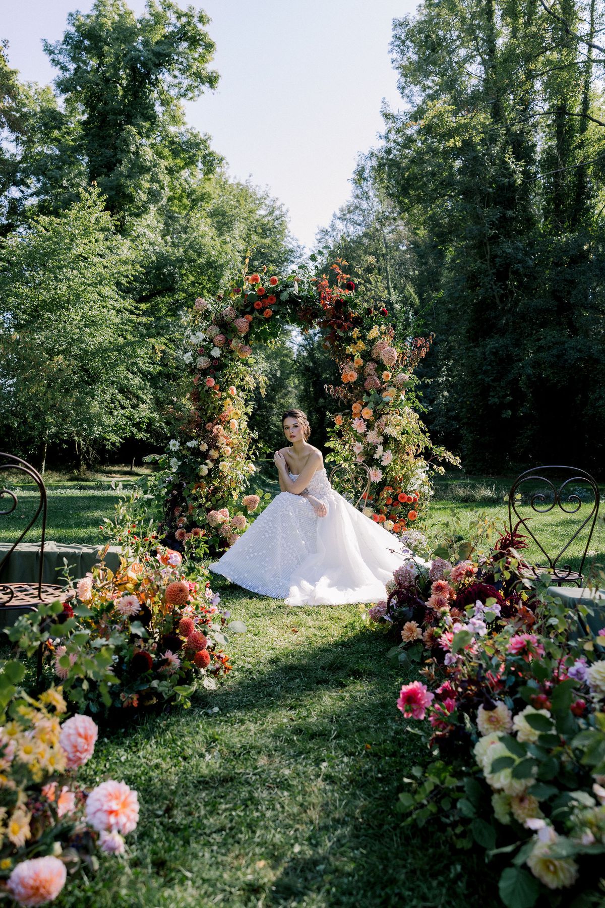 A wide view of a woodland garden with a tall floral arch, flower-lined lawn and a bride in a white gown in the distance.