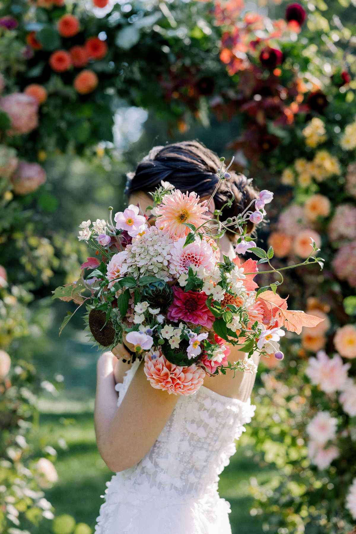 A bride with her hair pinned up looks away, holding a large pink and orange bouquet against a wall of flowers.