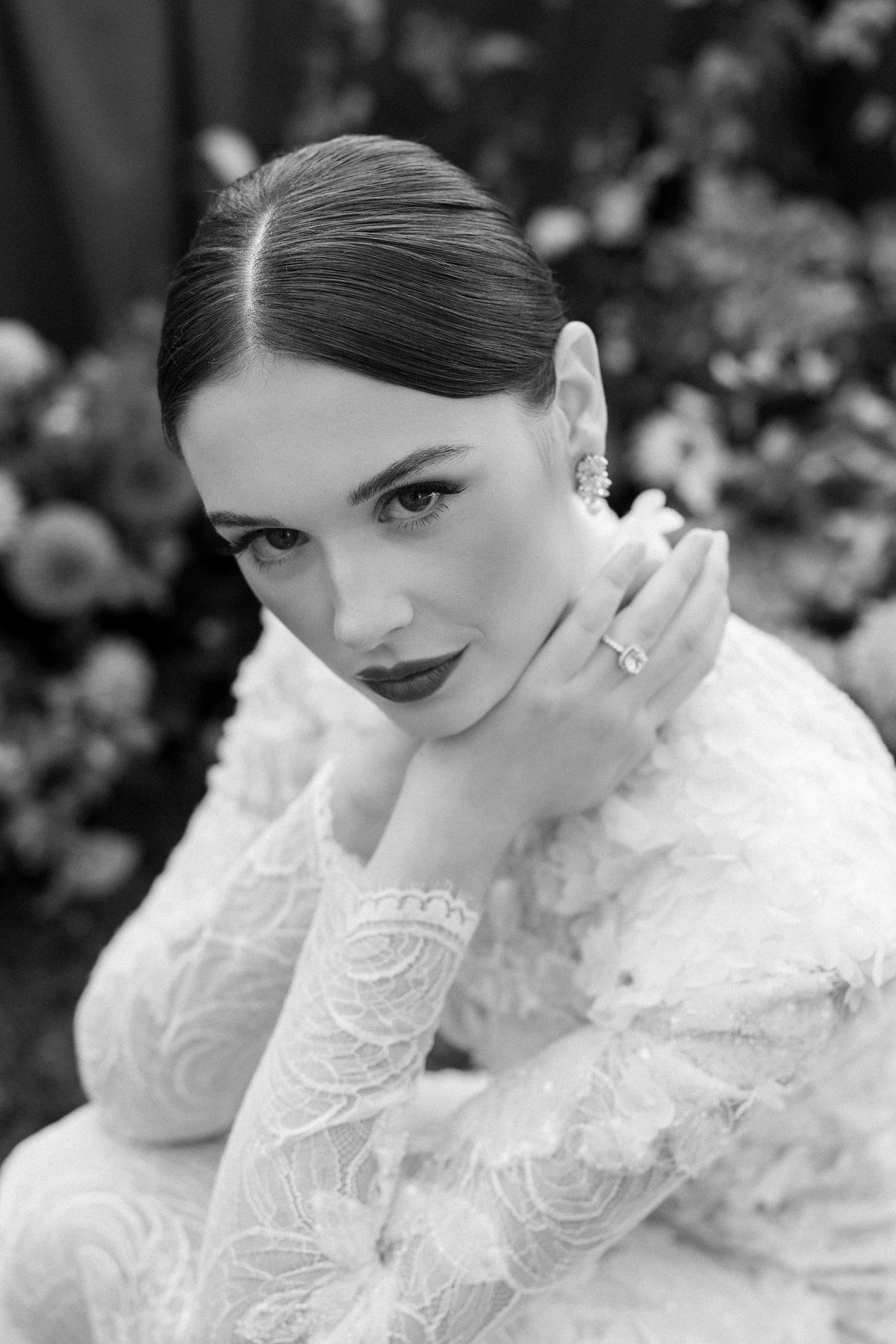 A black and white close portrait of the bride with centre-parted hair, dark lips and a ring, resting her chin on her hand.