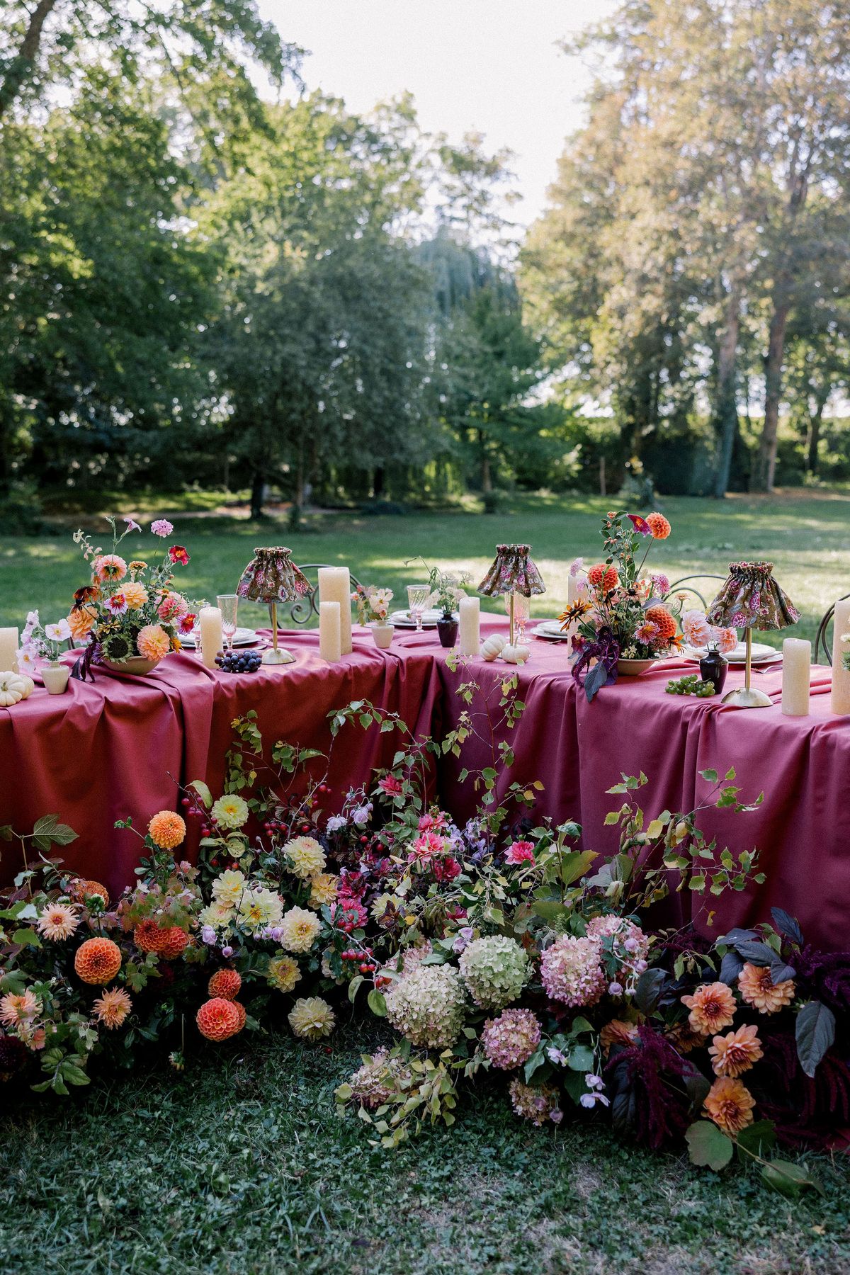 A banquet table with a raspberry tablecloth and lamps sits on grass, ringed by dense arrangements of colourful flowers among trees.