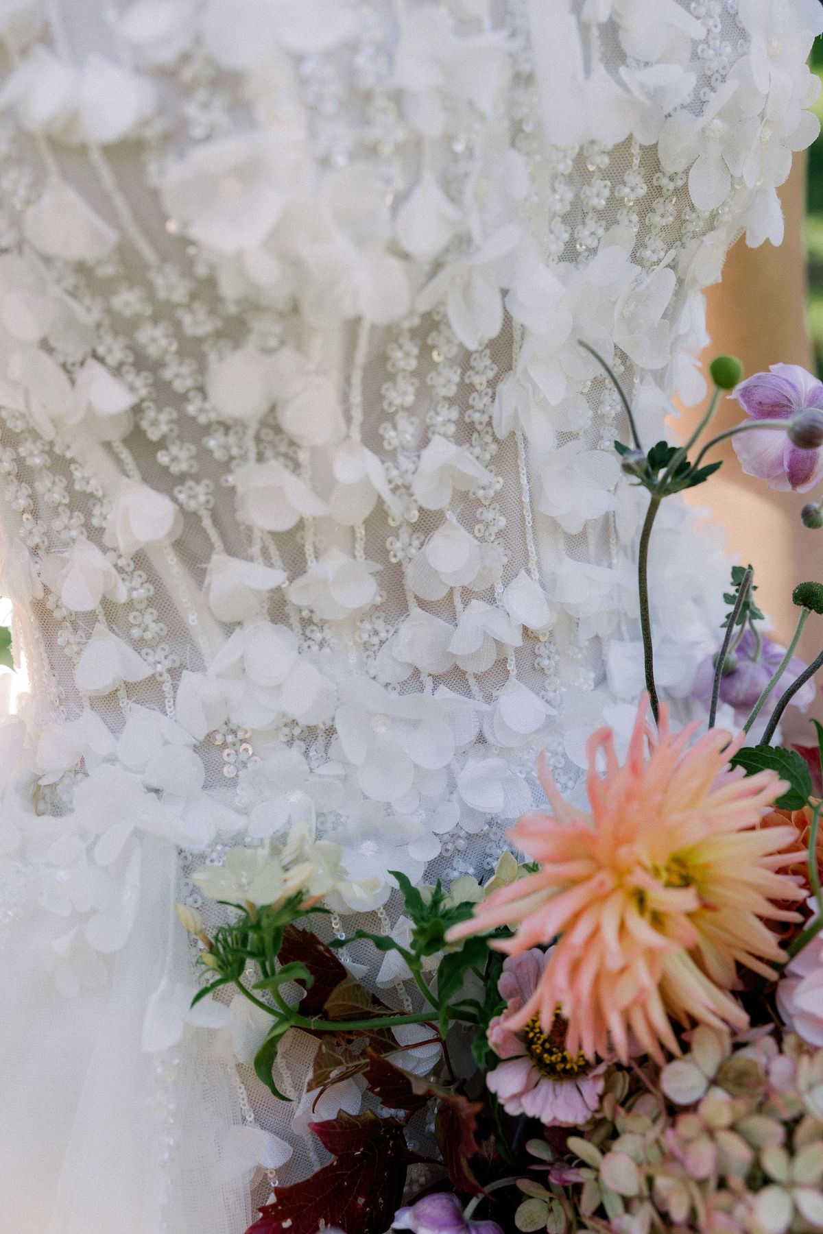 A close view of white floral appliqué on a gown beside an orange-pink dahlia and purple flowers.