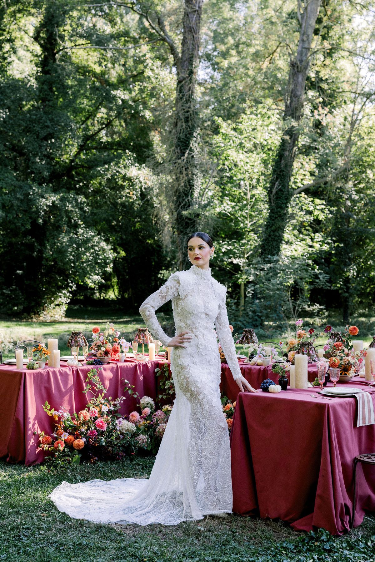 A bride in a long-sleeved lace gown stands with a hand on her hip beside a raspberry-covered table among tall trees.