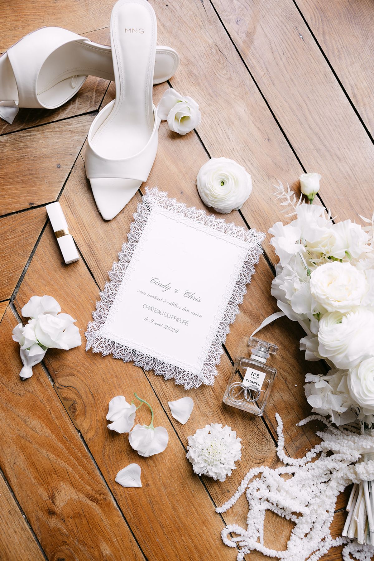 White heeled shoes, a framed invitation, a perfume bottle, a lipstick and white flowers are arranged on a wooden floor.