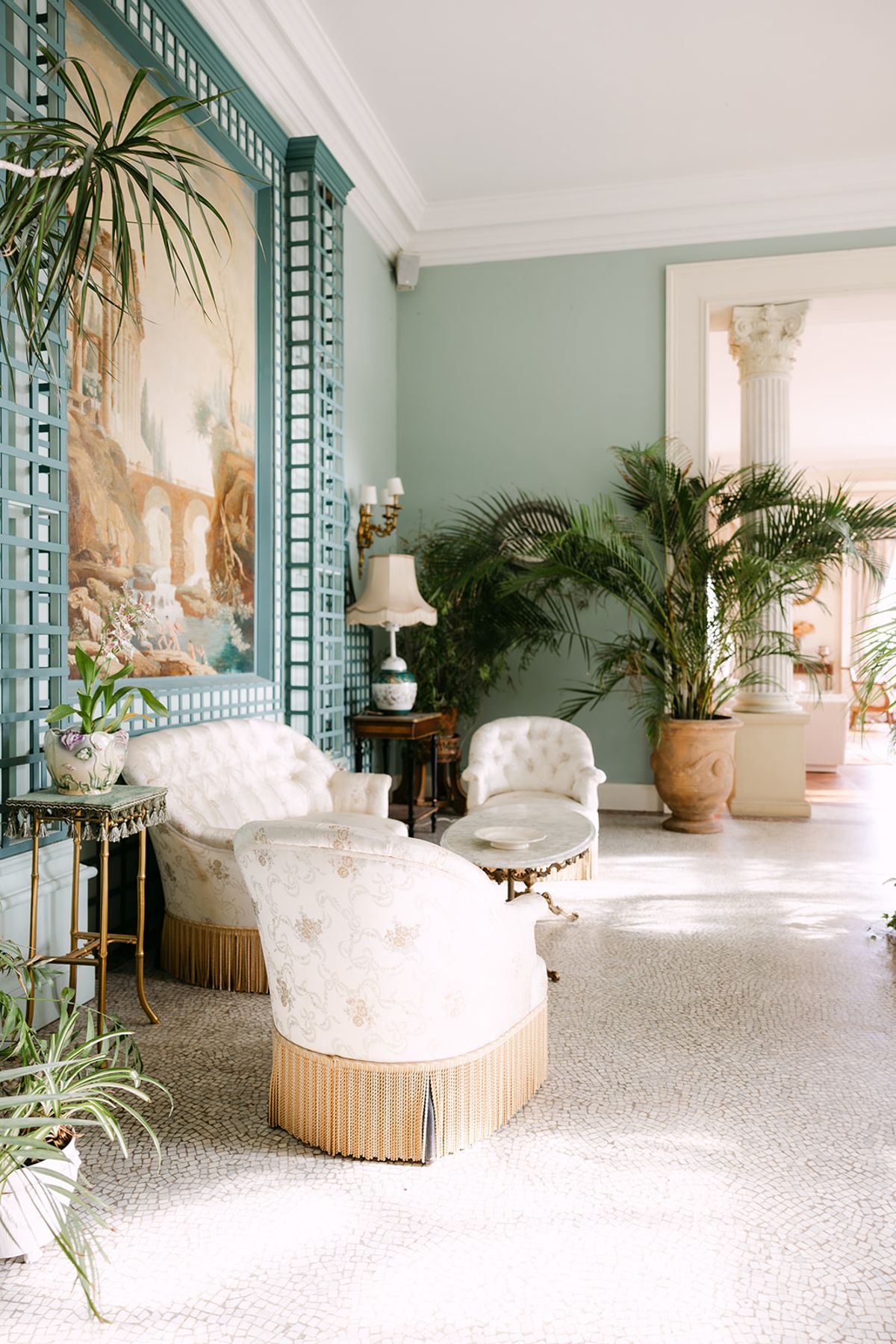 A bright room with pale green walls holds cream upholstered armchairs and potted palm plants, with light flooding in from the windows.