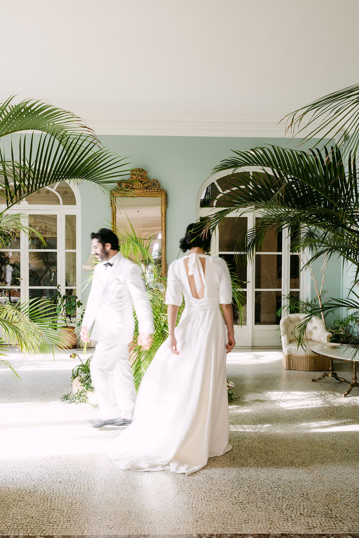 The couple, both in white, walk away from the camera through a sunlit conservatory with palm plants and tall arched windows.
