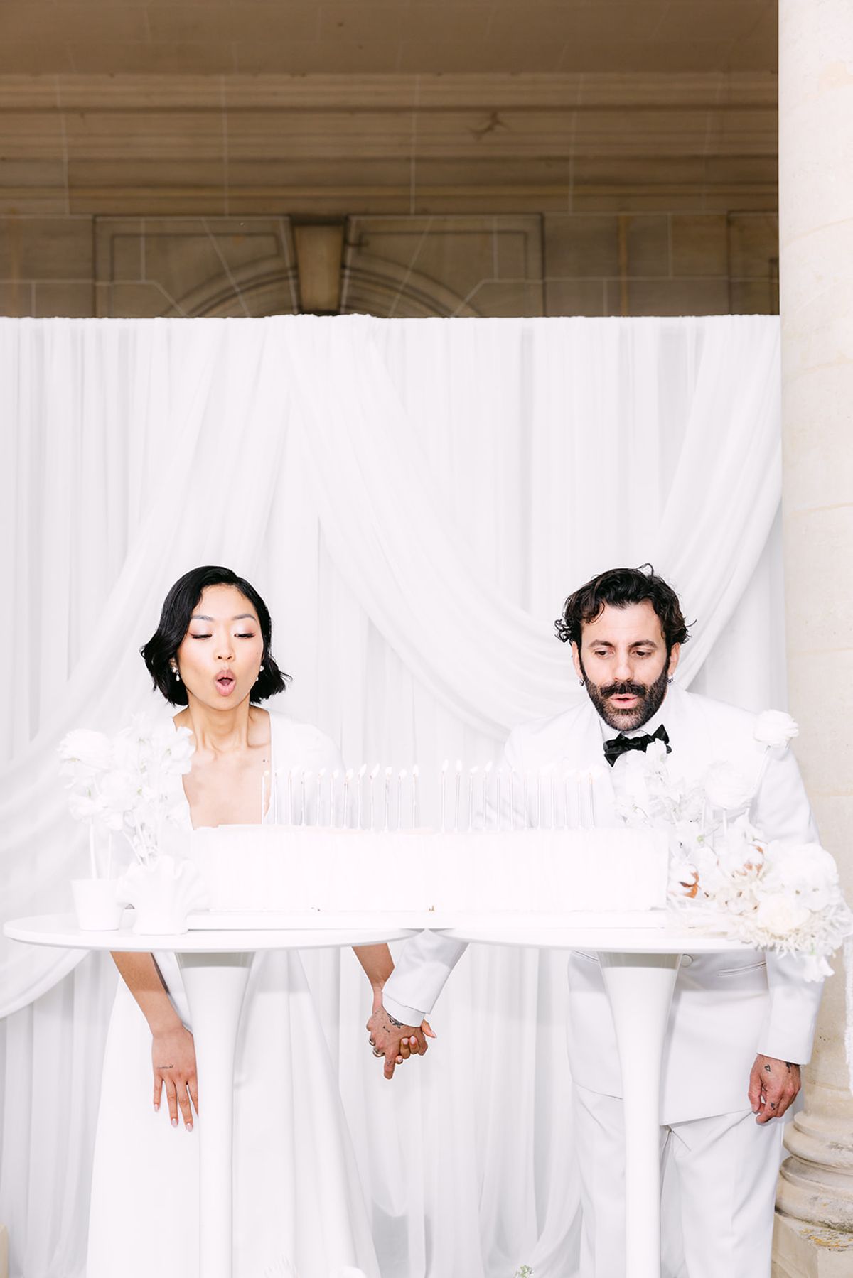 Bride and groom holding hands behind the long white cake, cheeks puffed to blow out the candles