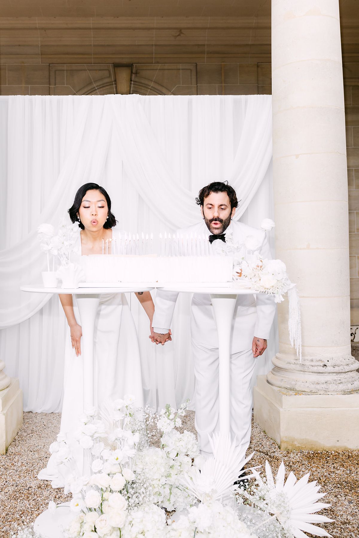 The couple stand behind a white table with flowers between stone columns, both leaning forward with pursed lips.