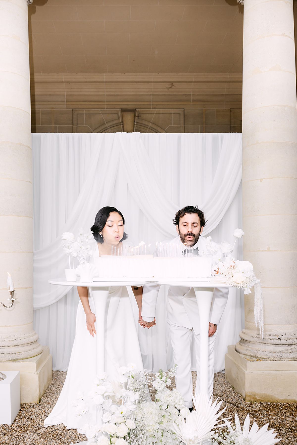 Bride and groom behind a long white cake on twin white tables, framed by stone columns and draped fabric