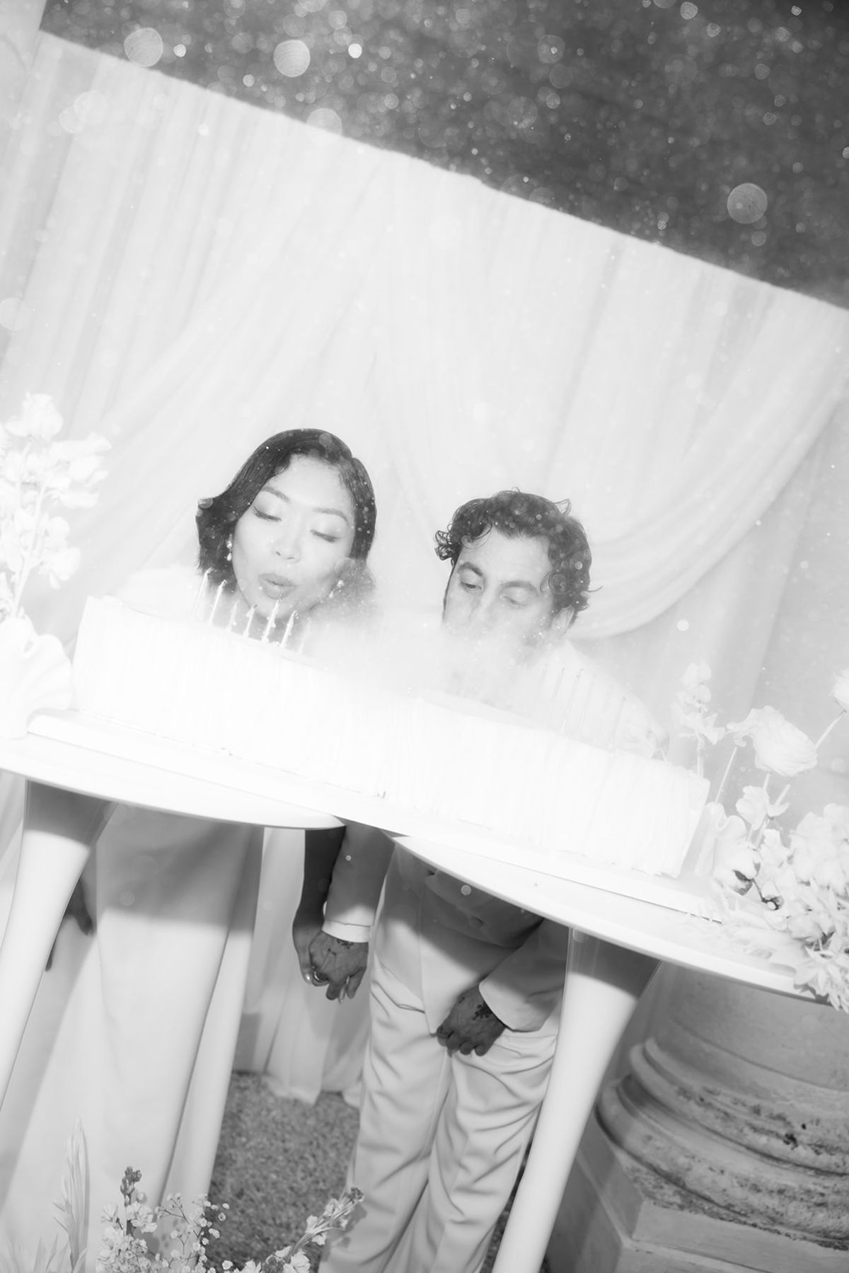 In black and white, the couple lean over a white cake, blowing towards it, with white drapes behind them.