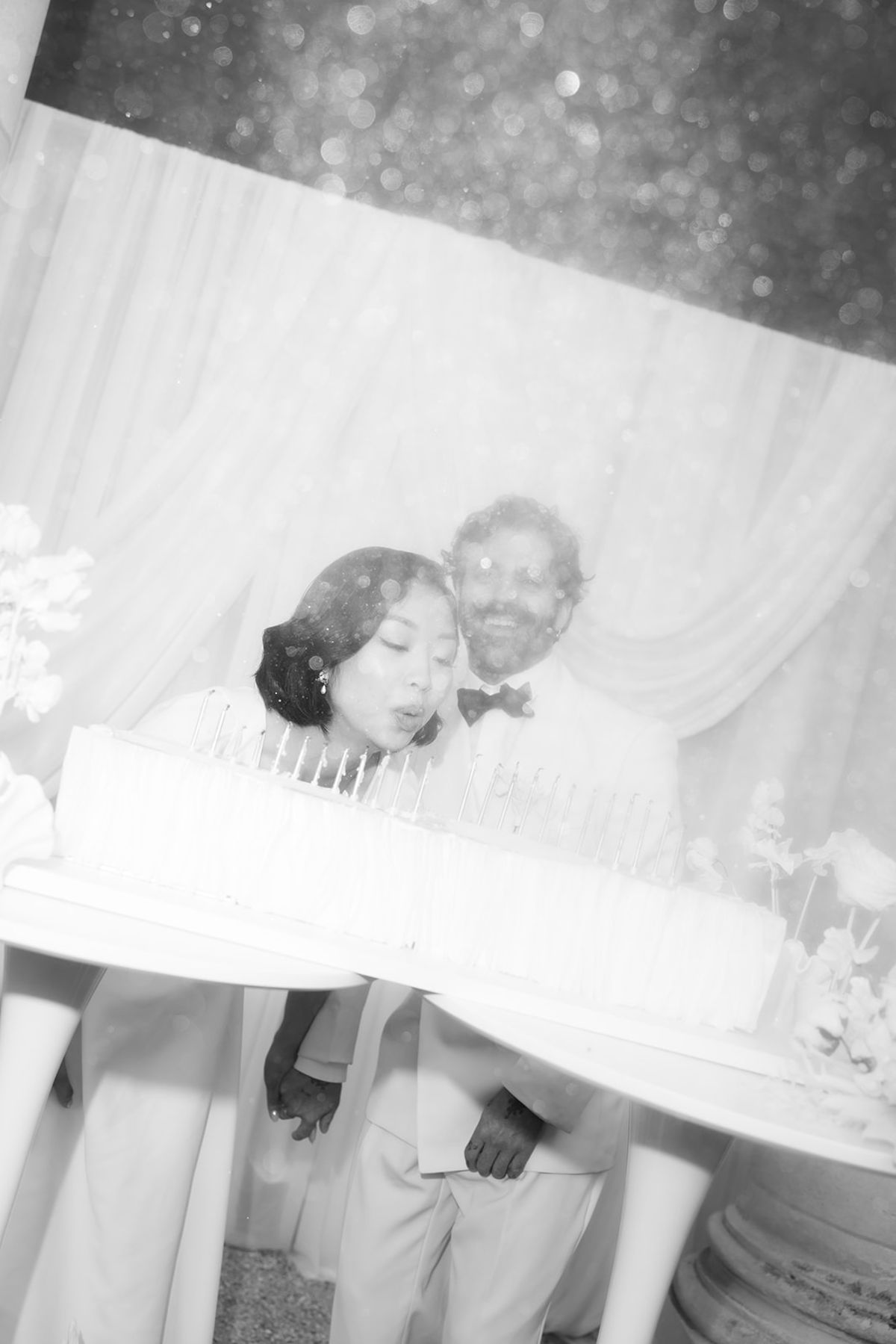 Black and white frame of the bride and groom leaning over the cake to blow out the candles