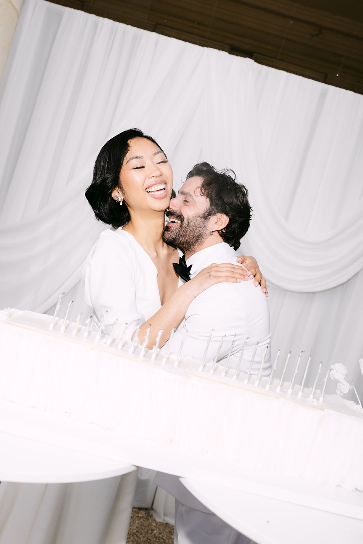 The bride and groom laugh as they embrace in front of white draped fabric, with glasses lined up in the foreground.