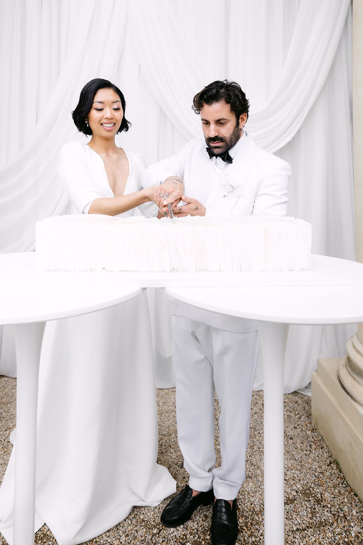 The bride and groom stand behind a large white table, both reaching towards a white display of glasses as if cutting a cake.