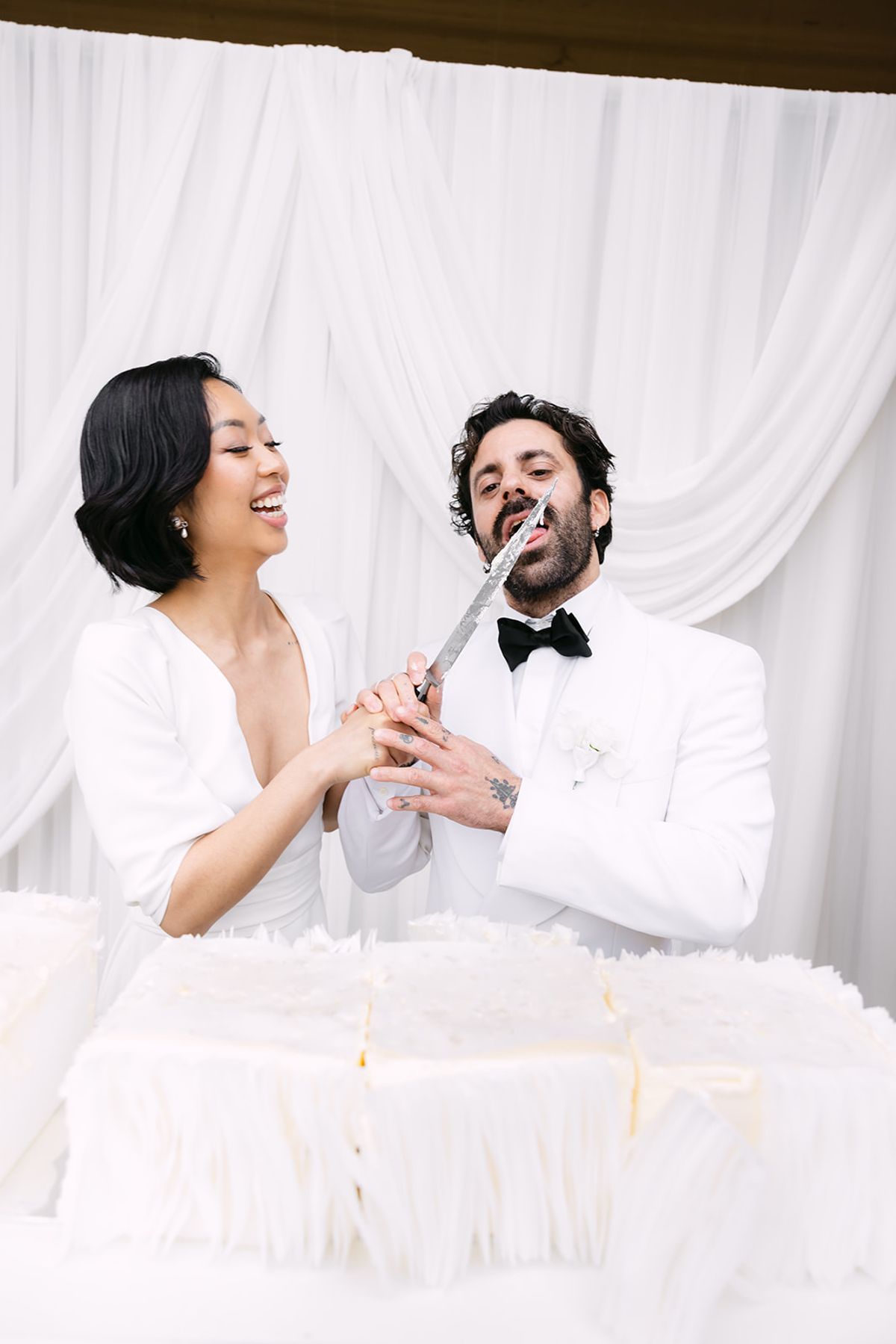 The bride laughs as she and the groom hold a knife over a white cake, both dressed in white before drapes.