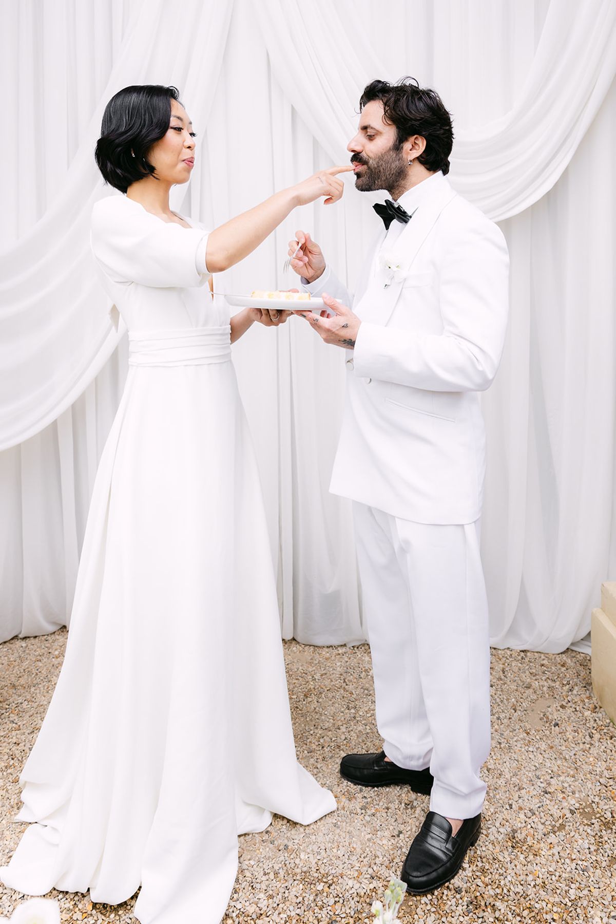 The bride in a long white dress holds out a fork to the groom, who leans in to taste a bite of cake.
